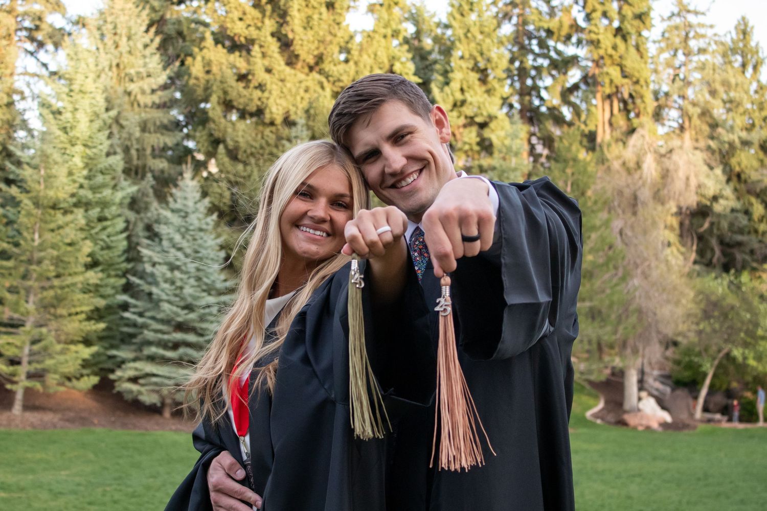 Two graduates in black robes smile and point at camera outdoors with evergreen trees in background.