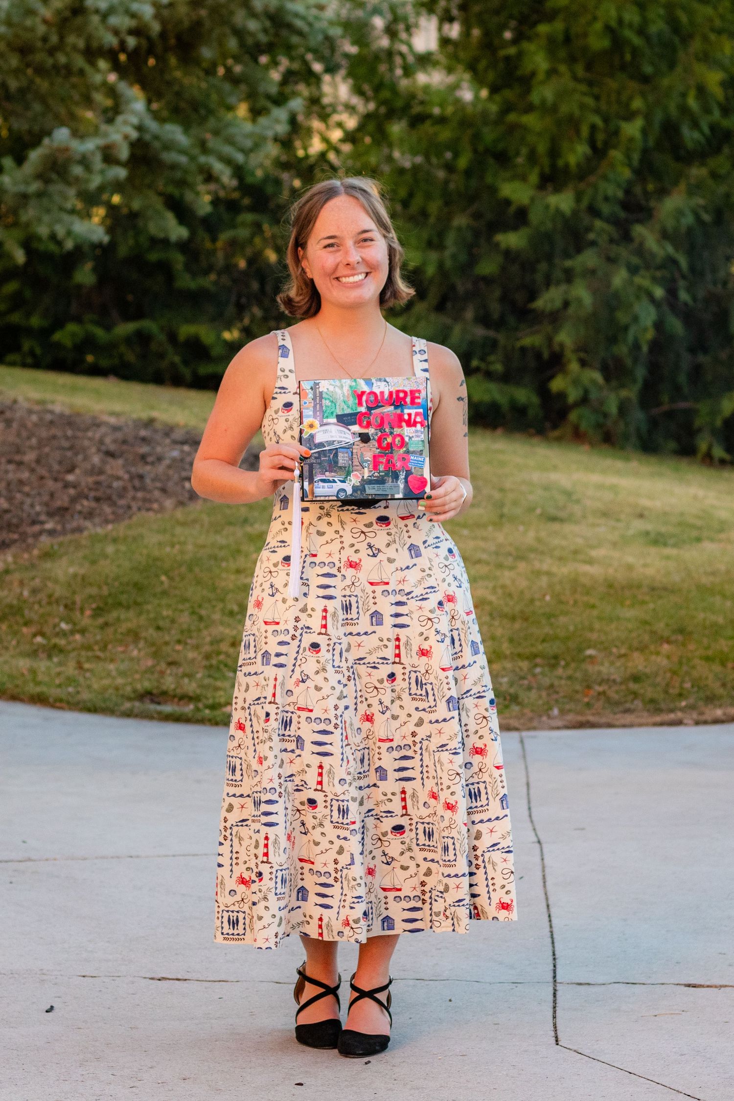 Smiling woman in printed midi dress holding colorful book outdoors on sunny day.