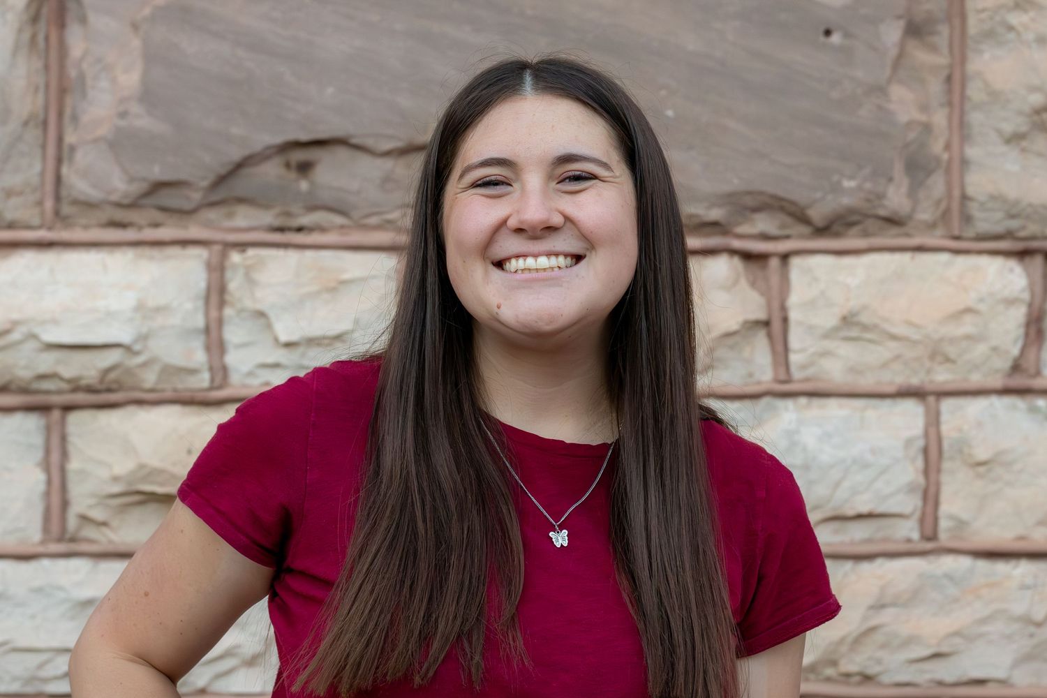 Smiling young woman with long brown hair in red t-shirt poses in front of stone wall.