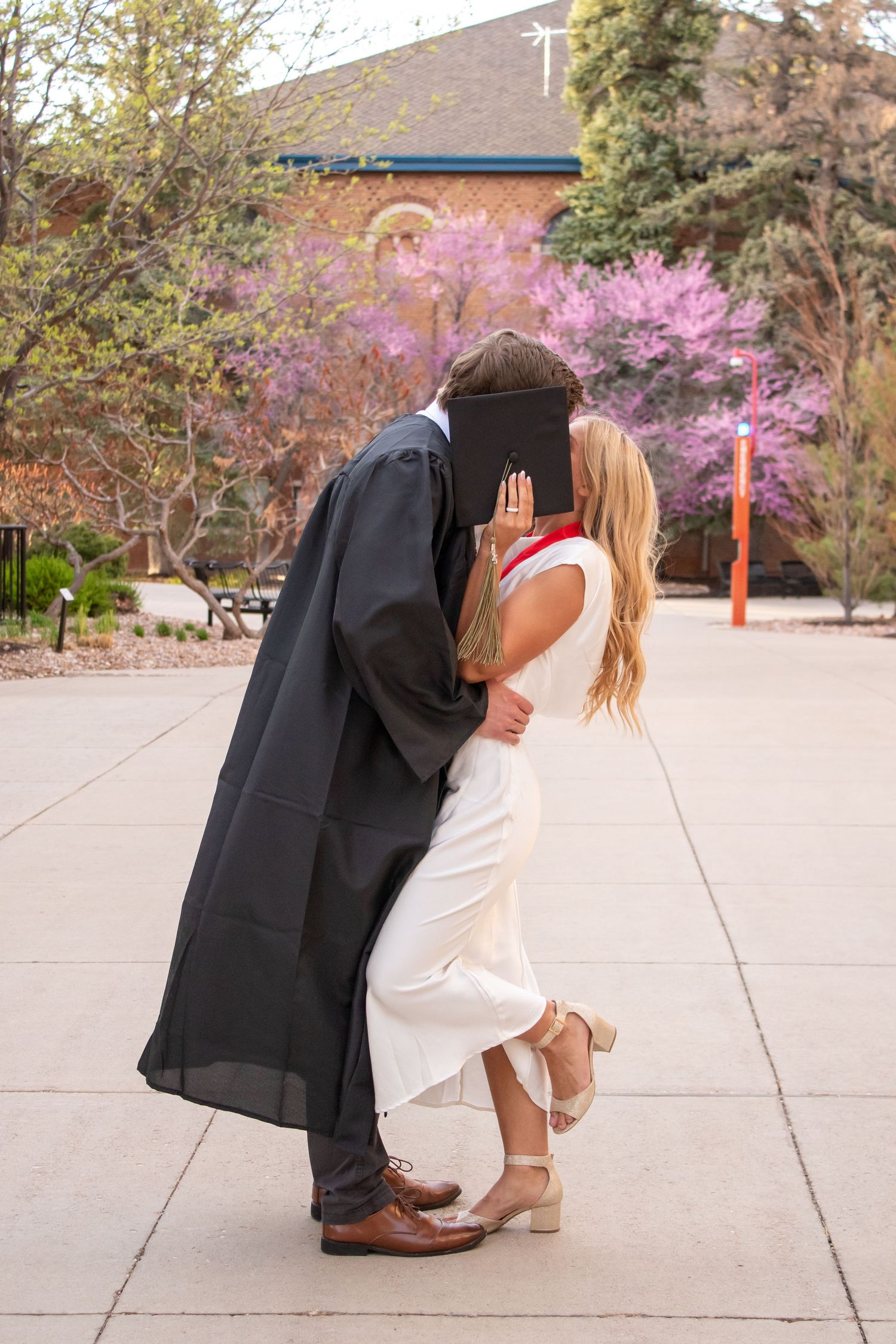 Couple sharing a kiss at graduation, man in black gown holding cap, woman in white dress, spring trees in background.