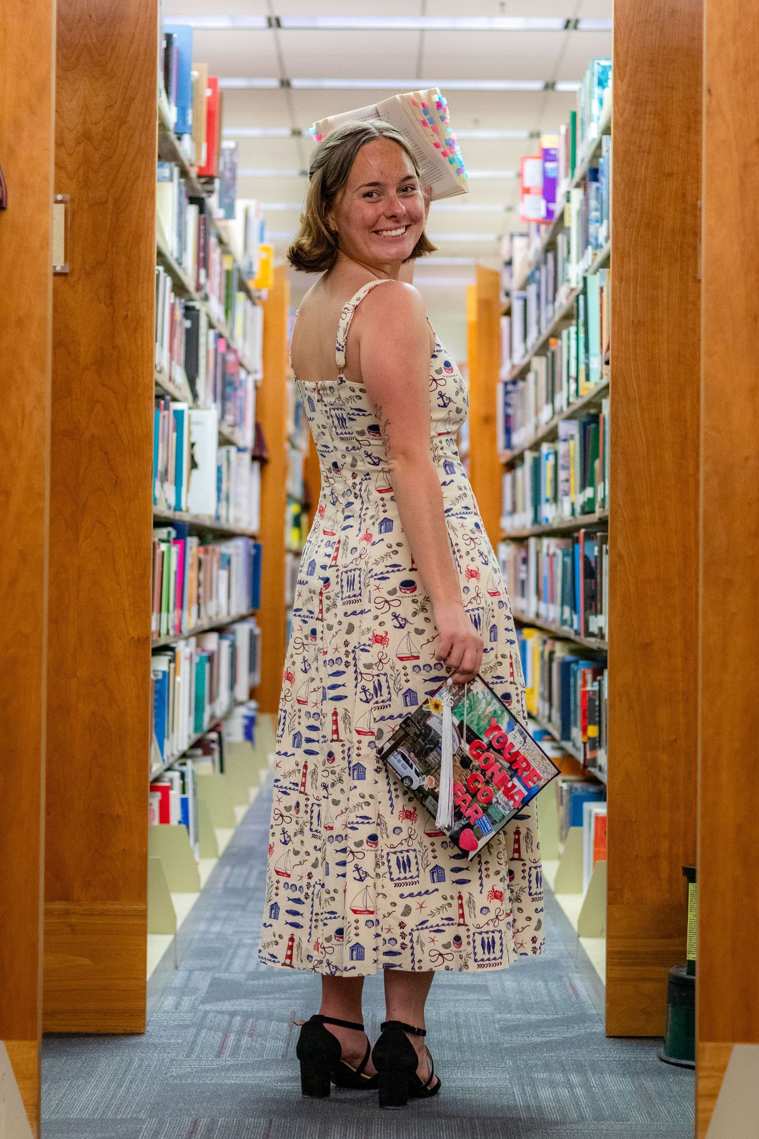 Smiling woman in floral dress holding a book between library shelves.