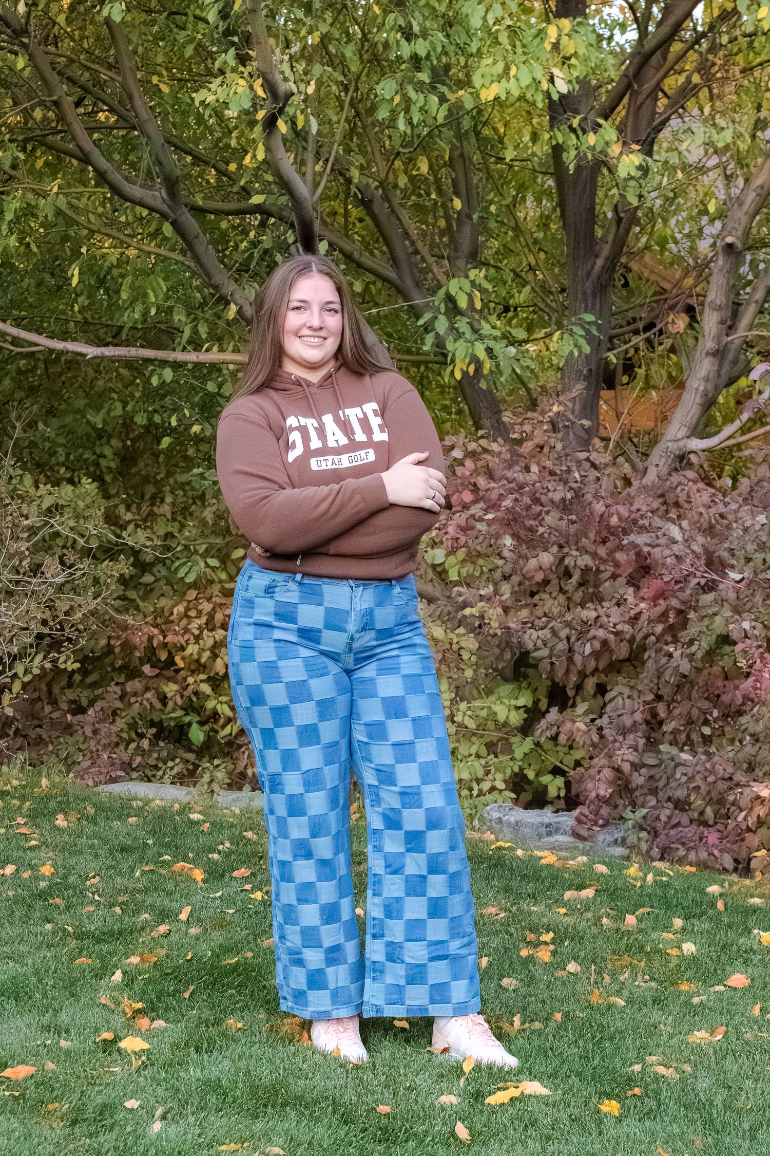 Cheerful young woman in brown State hoodie and blue checkered jeans posing with arms crossed outdoors.