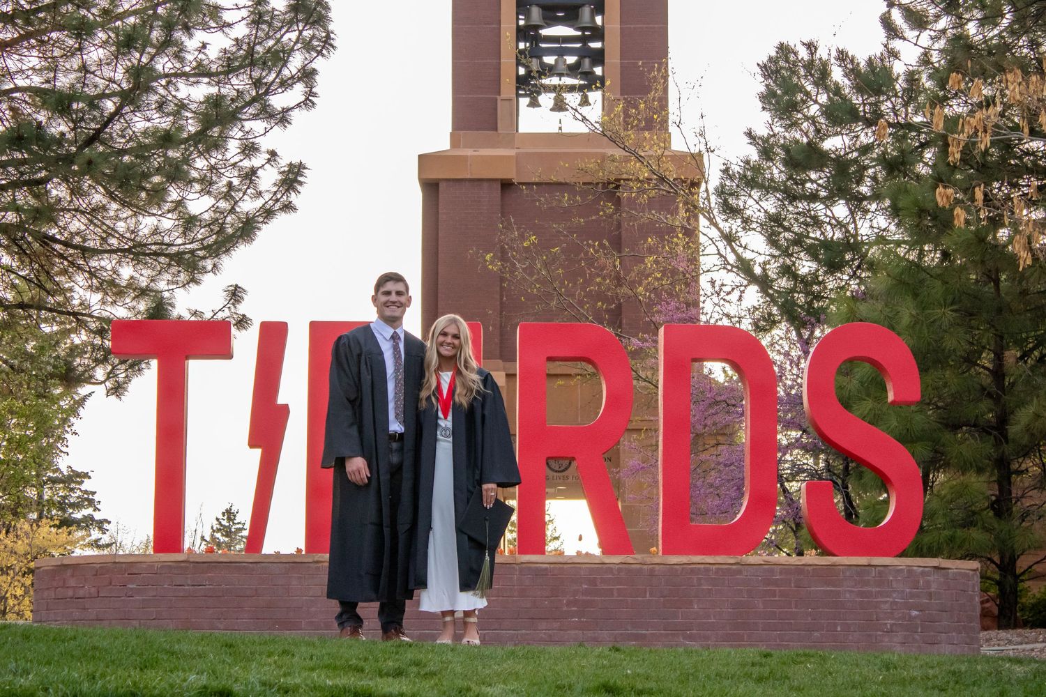 Two graduates in caps and gowns pose in front of red THUNDERBIRDS sign with bell tower at university campus.
