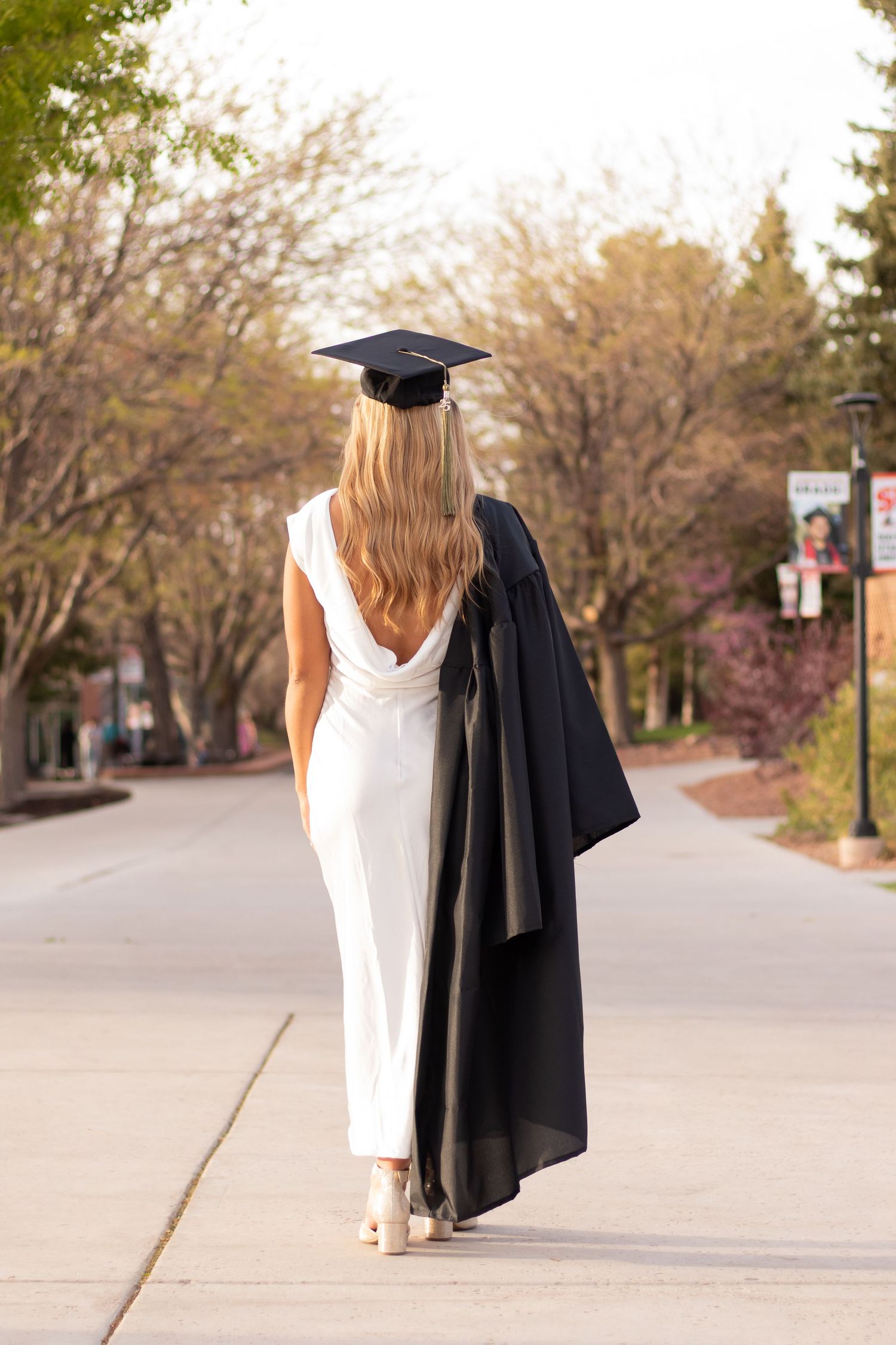 A blonde woman in a white dress and graduation cap walks away, holding her black gown on a tree-lined campus path.