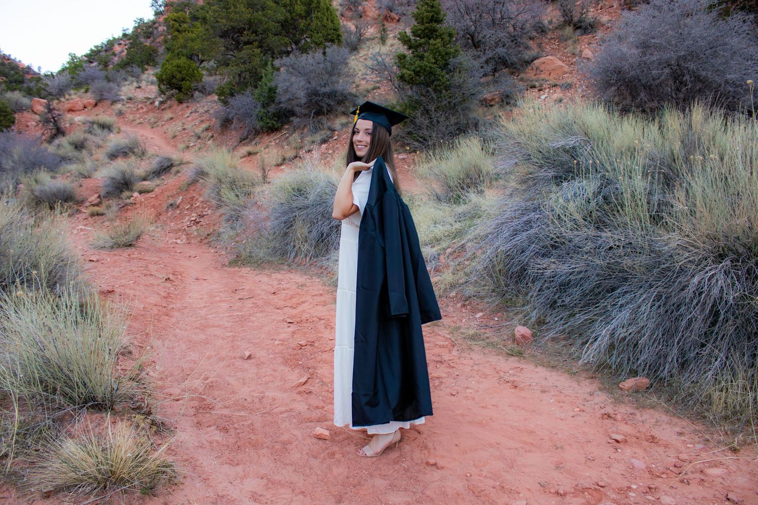 Young woman in black graduation cap and gown over white dress poses on red dirt path in desert landscape.