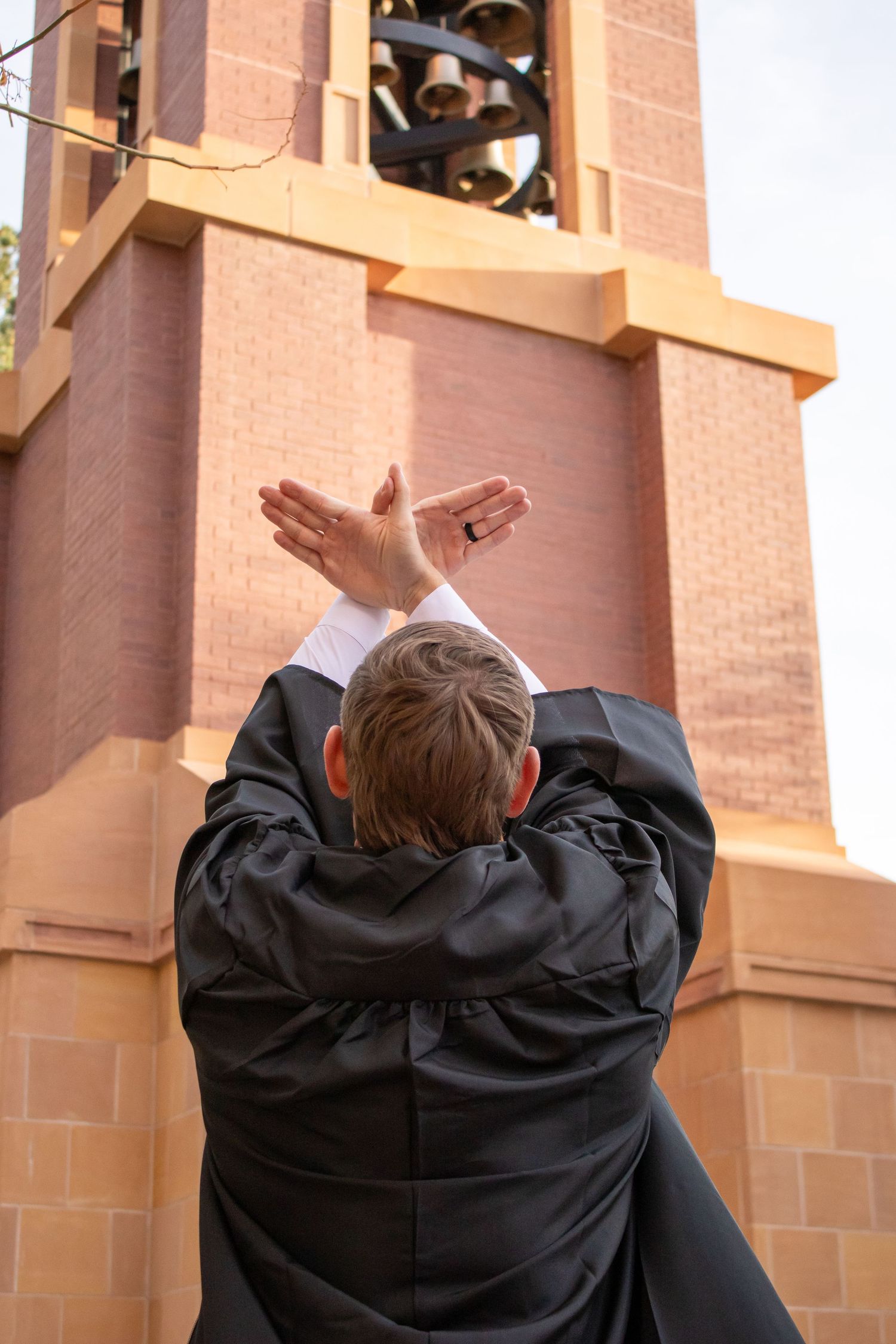 A robed clergy member raises hands toward a brick church bell tower in an outdoor blessing gesture.