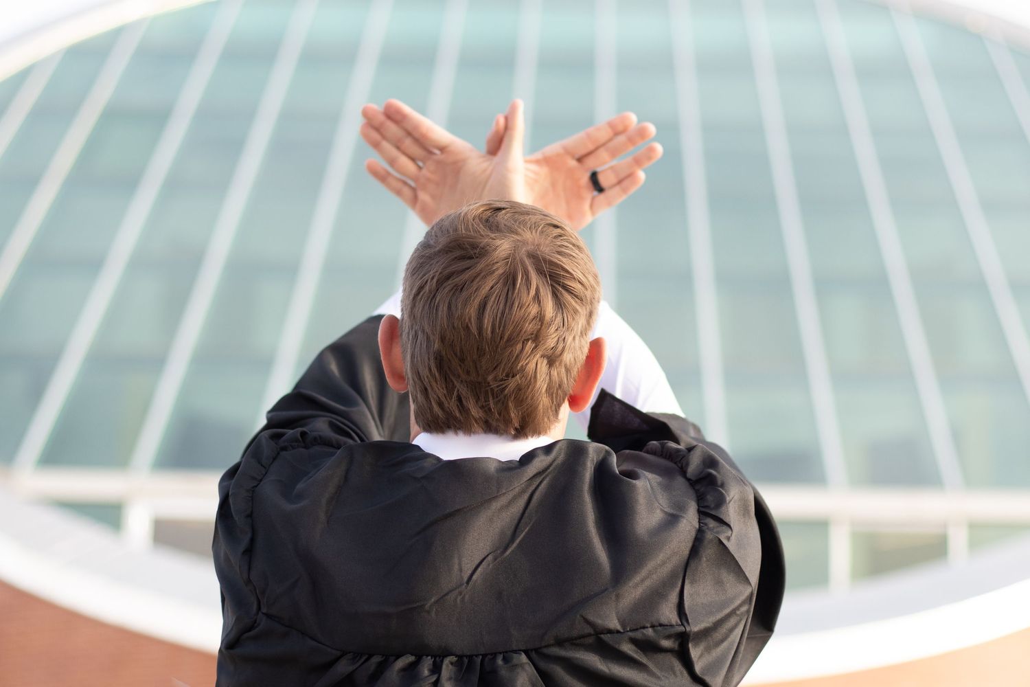 Man in black jacket making bunny ears gesture with hands behind his head, standing outdoors near a modern building.