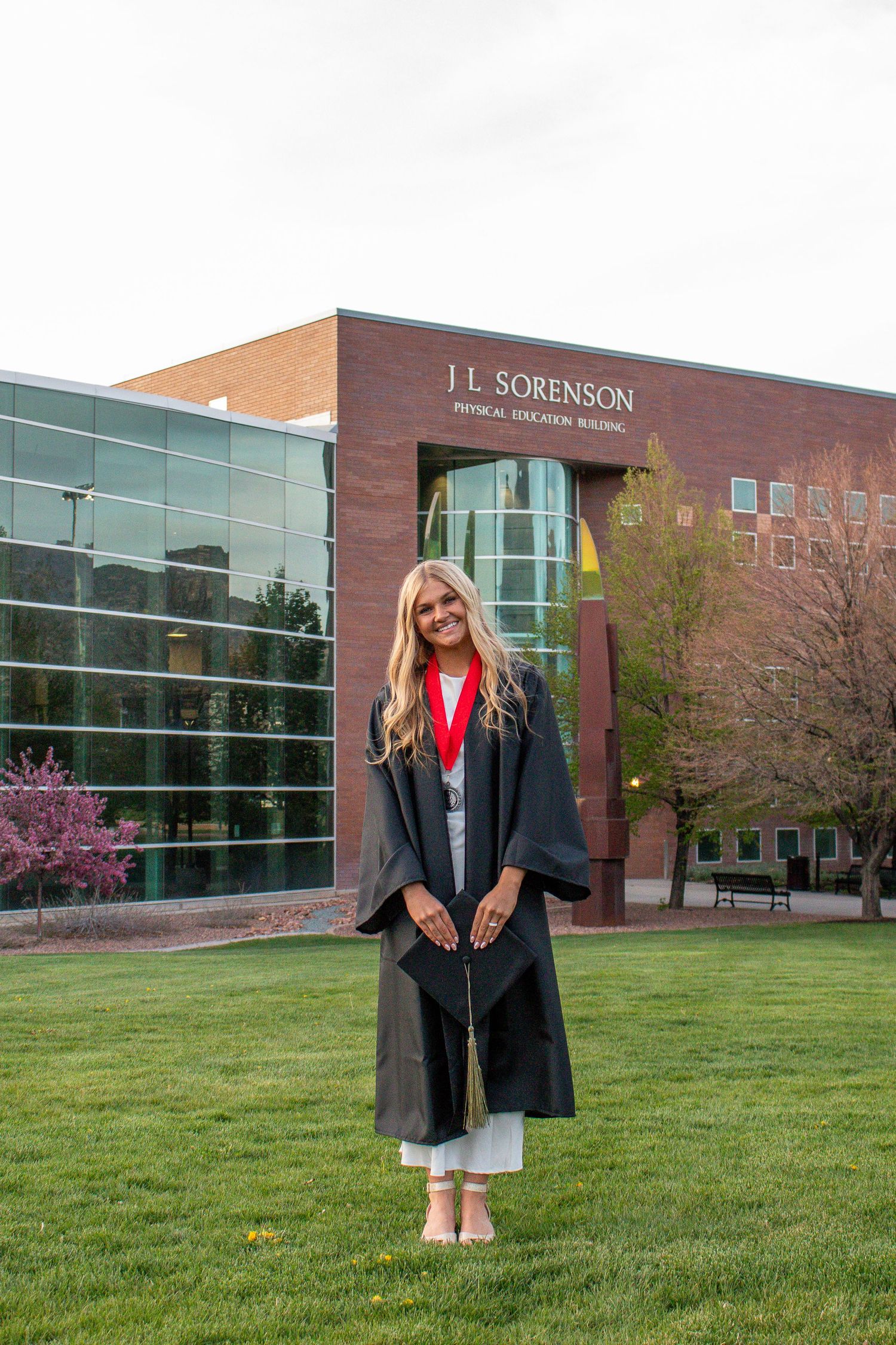 A smiling graduate in cap and gown stands on green lawn before the J.L. Sorenson Physical Education Building.
