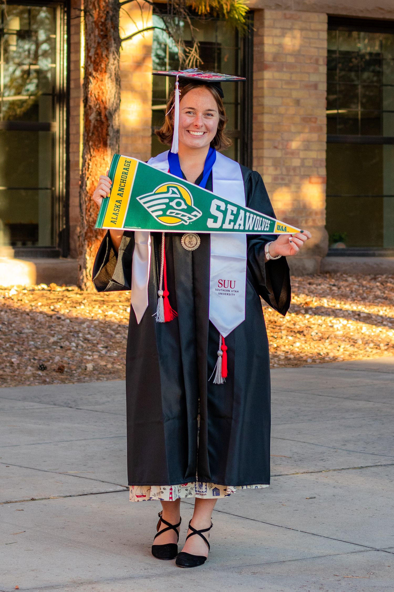 Graduate in cap and gown holds Alaska Anchorage Seawolves pennant and SUU stole, smiling outdoors on campus.
