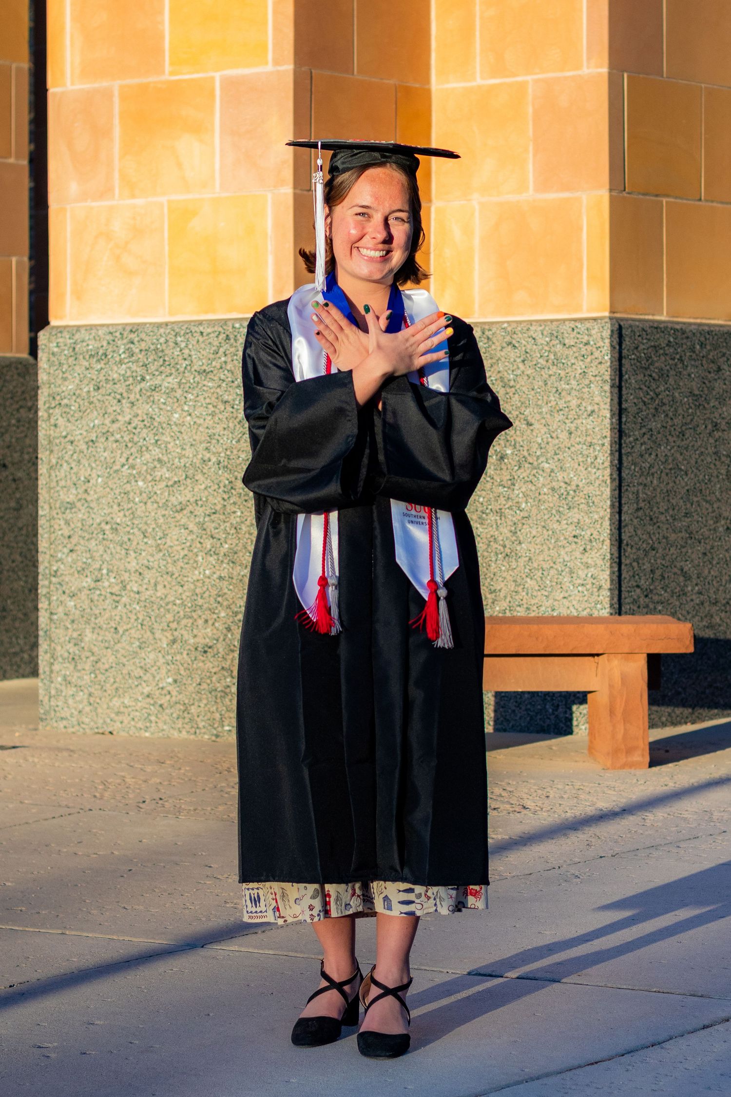 Smiling graduate in black cap and gown with colorful stoles poses proudly outdoors near stone building columns. Southern Utah University Tbi