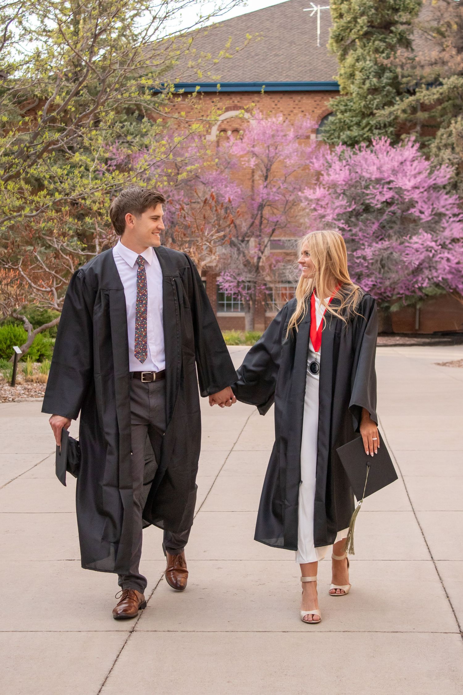 Couple in black graduation gowns holding hands and walking on campus pathway with blooming pink trees in background.