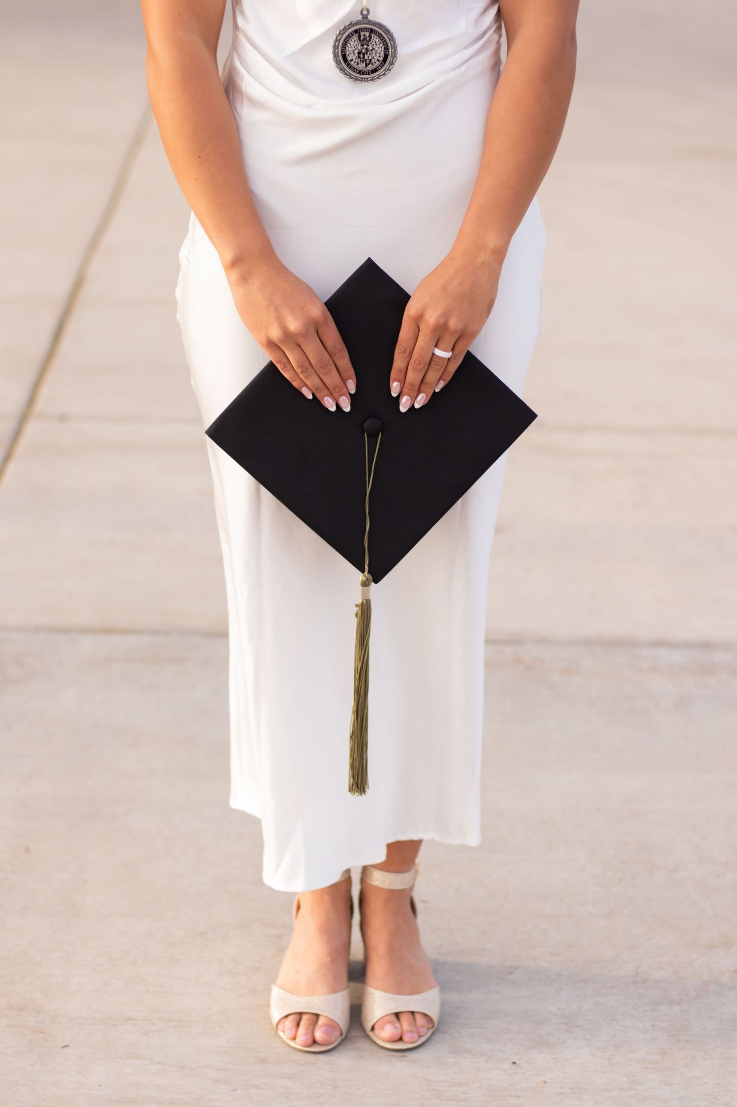 Graduate in white dress and heels holding black mortarboard cap with gold tassel, showcasing manicured nails.