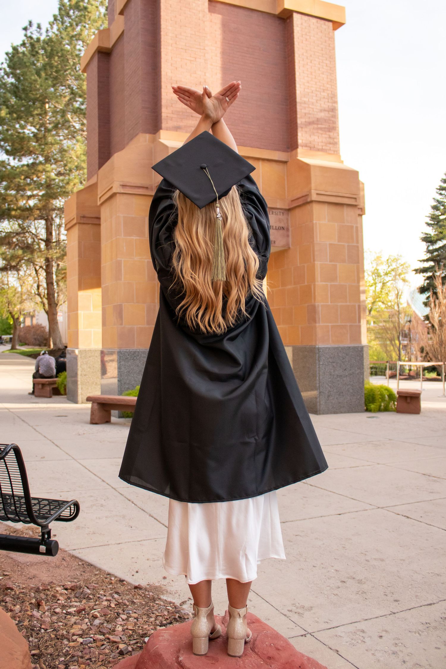 Female graduate in black cap and gown poses with arms raised at university campus outdoor ceremony.