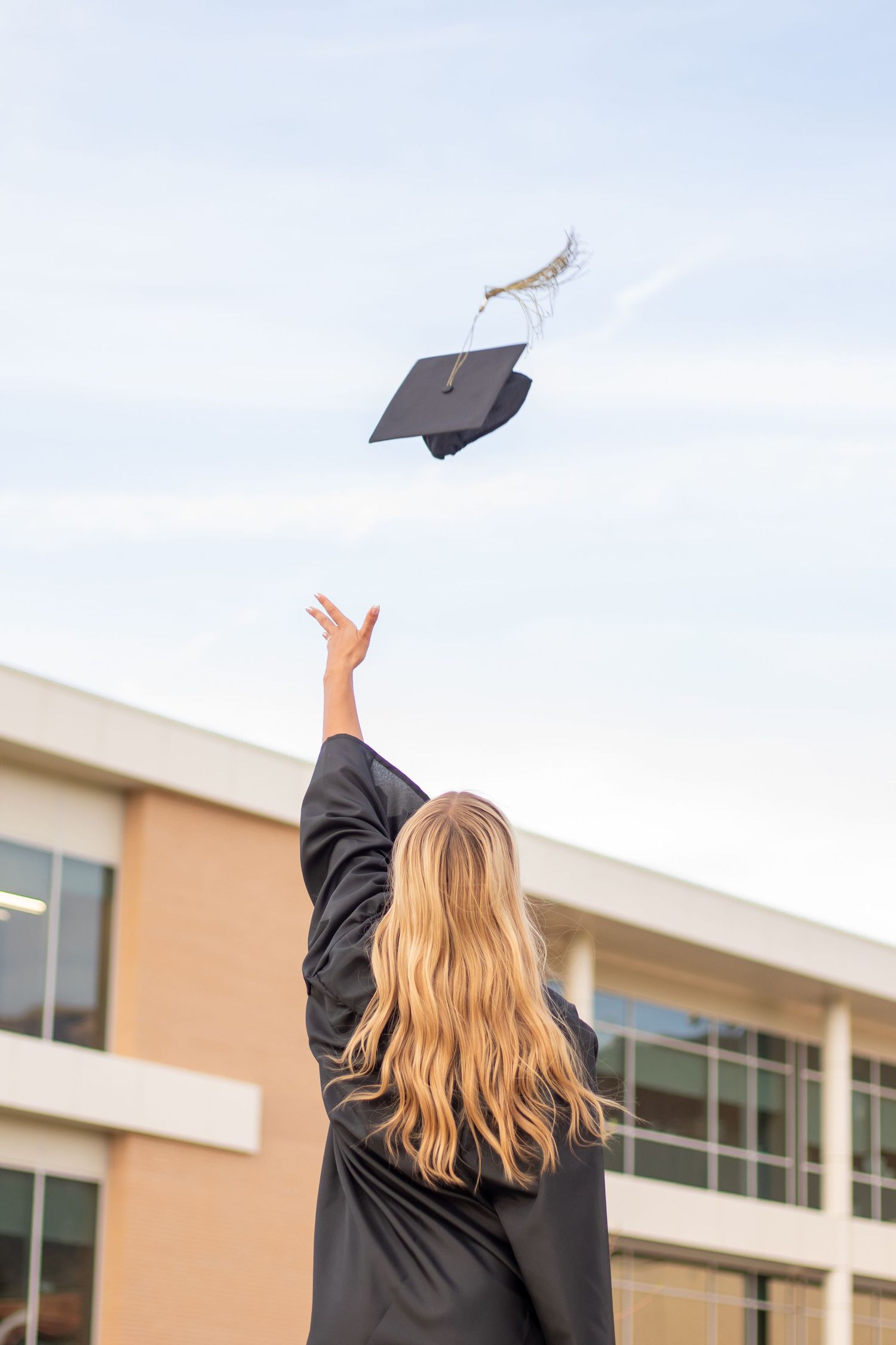 Graduate in black gown tossing mortarboard cap into blue sky, celebrating academic achievement outdoors near campus building.