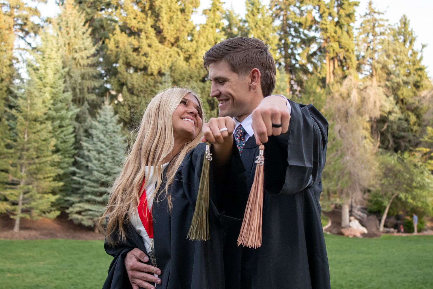 Two graduates in black gowns holding tassels and smiling at each other outdoors among tall green trees.