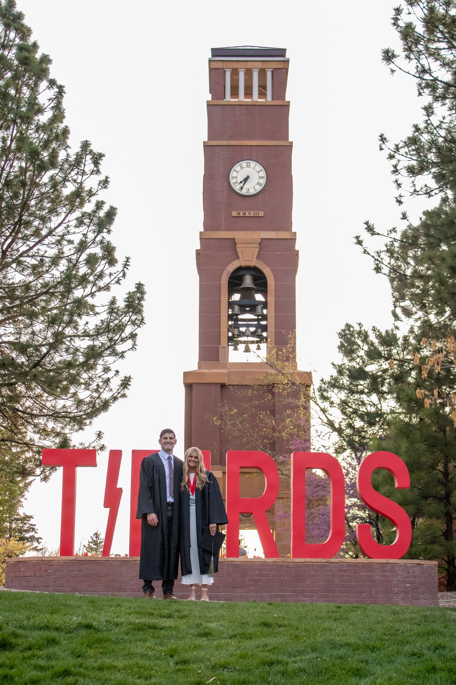 Two graduates in caps and gowns pose by a brick clock tower and red Thunderbirds sign on campus.