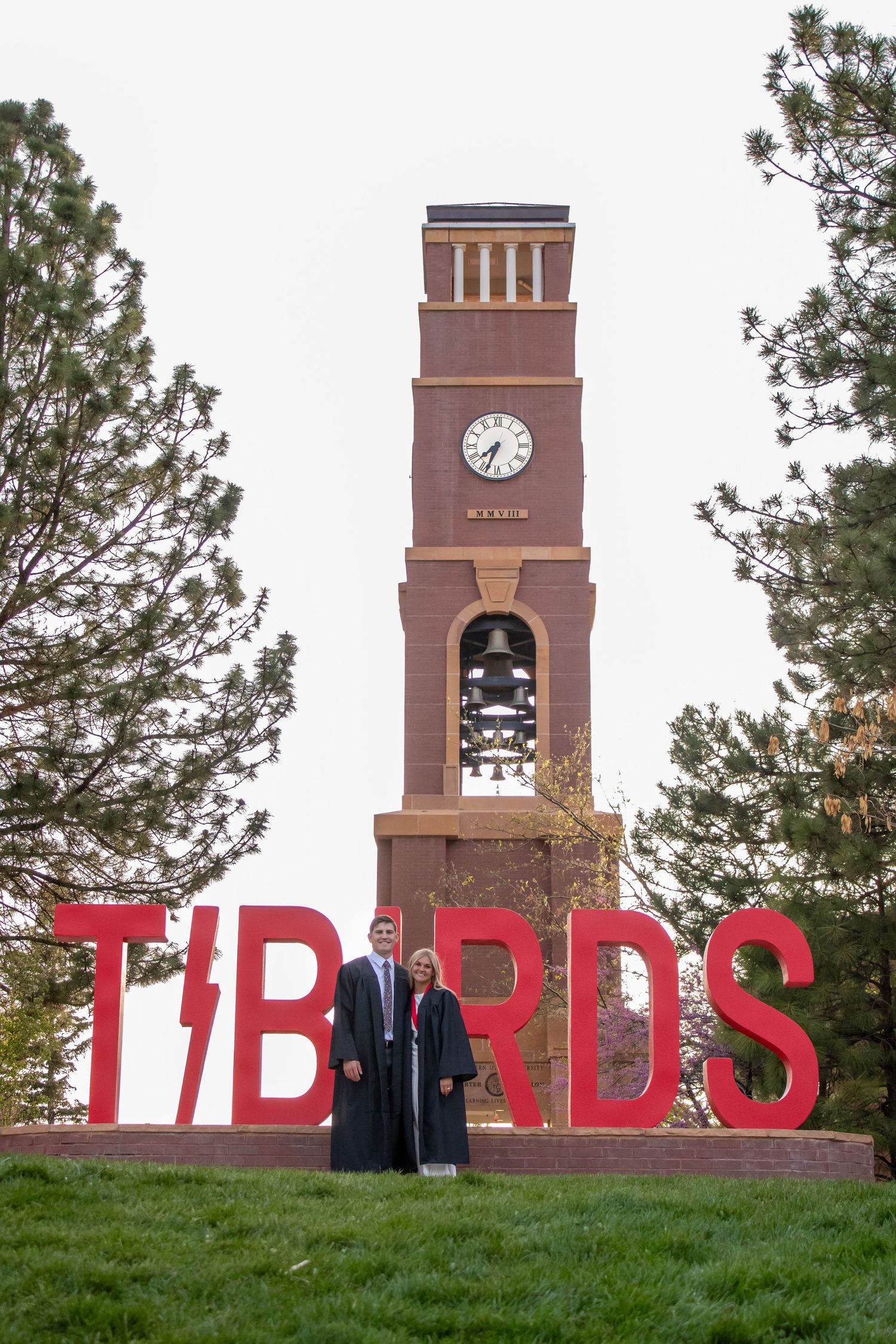 Two graduates in black gowns pose before a TIBIRDS sign and brick clock tower on a university campus.