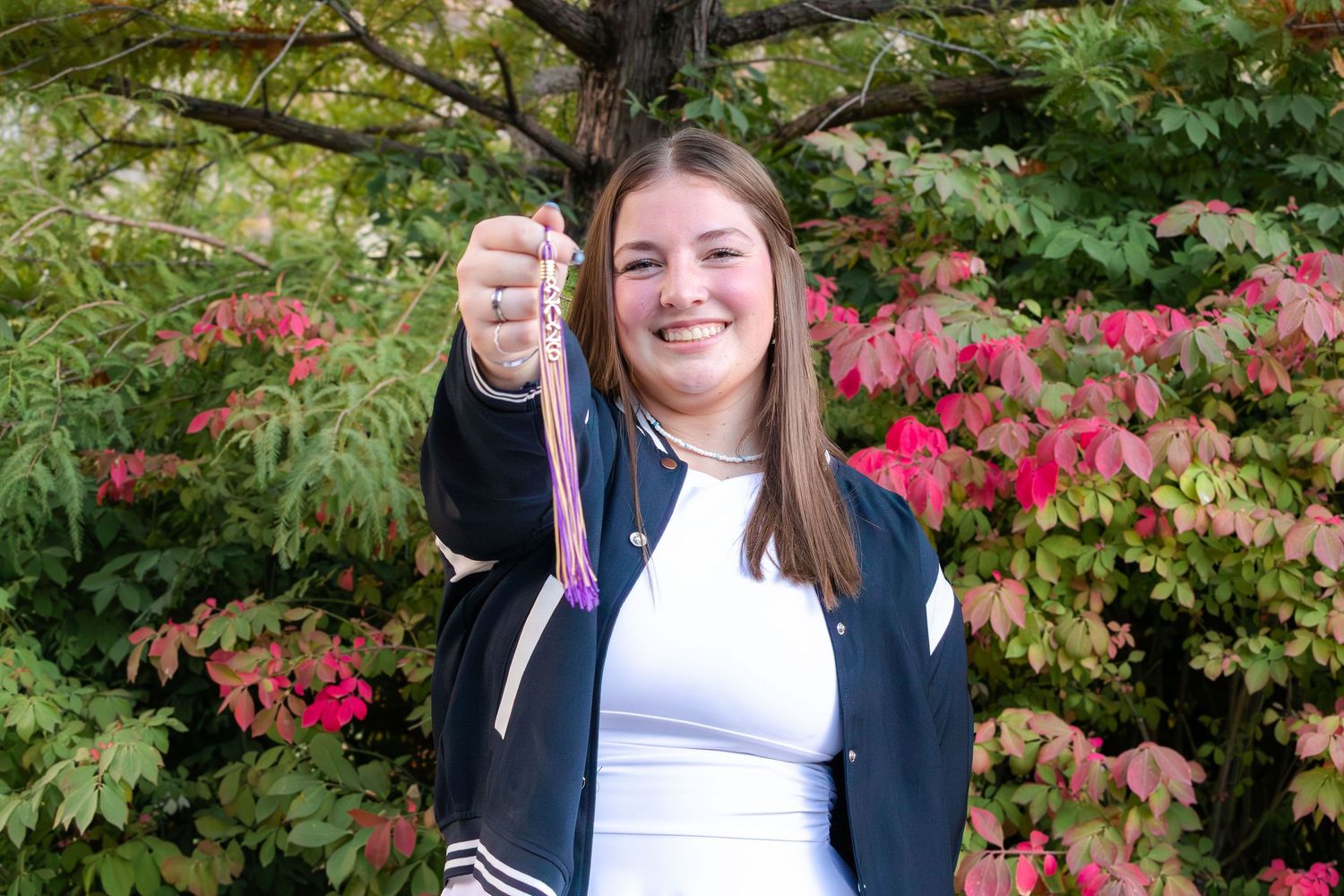 Smiling young woman holding jewelry while standing near vibrant red and pink autumn foliage outdoors.