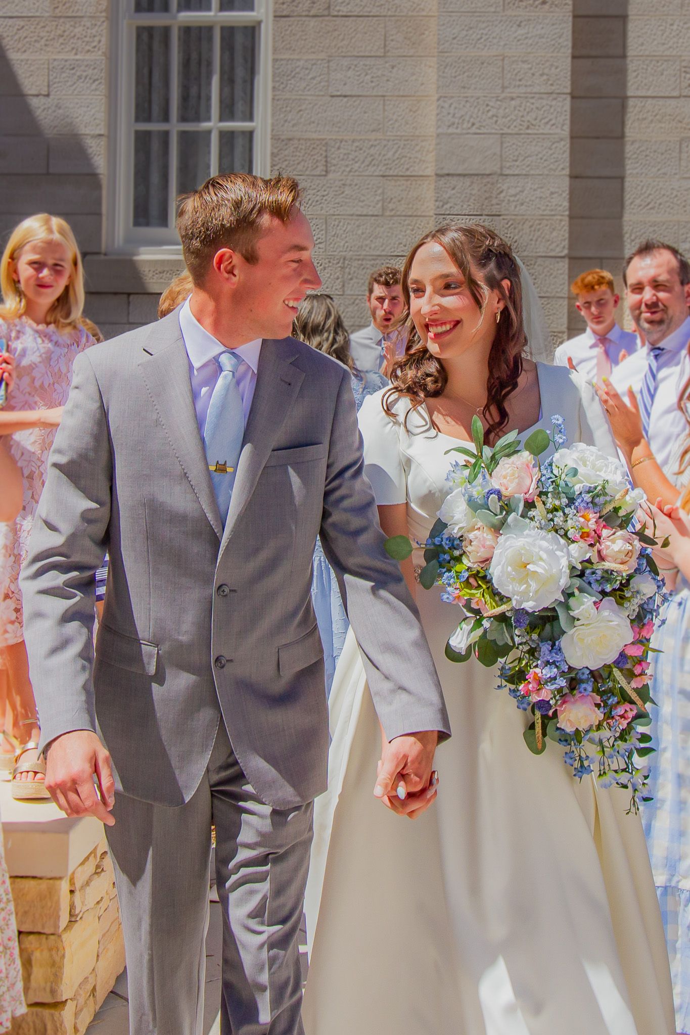 Happy bride and groom exit ceremony holding colorful bouquet among cheering guests.