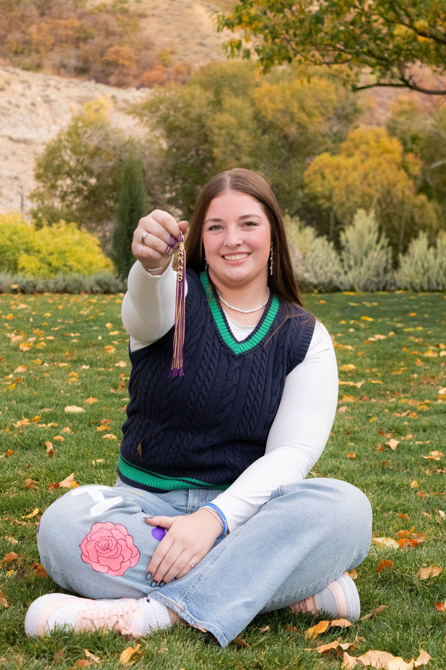 Smiling teen girl sitting cross-legged on grass holding a tassel, wearing jeans and navy vest in autumn park.