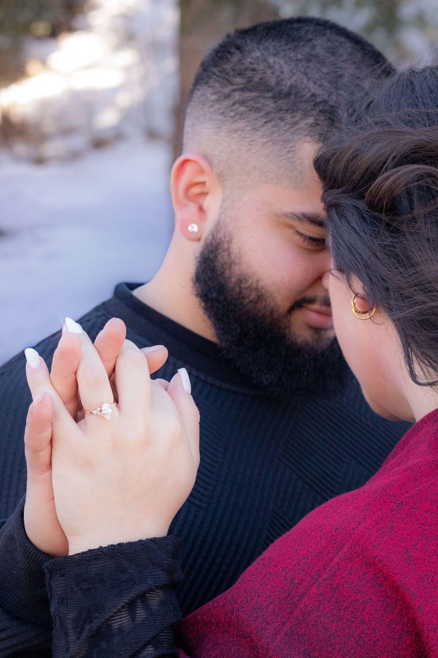 Engaged couple touching foreheads intimately in snowy setting, woman displaying diamond engagement ring while holding hands.