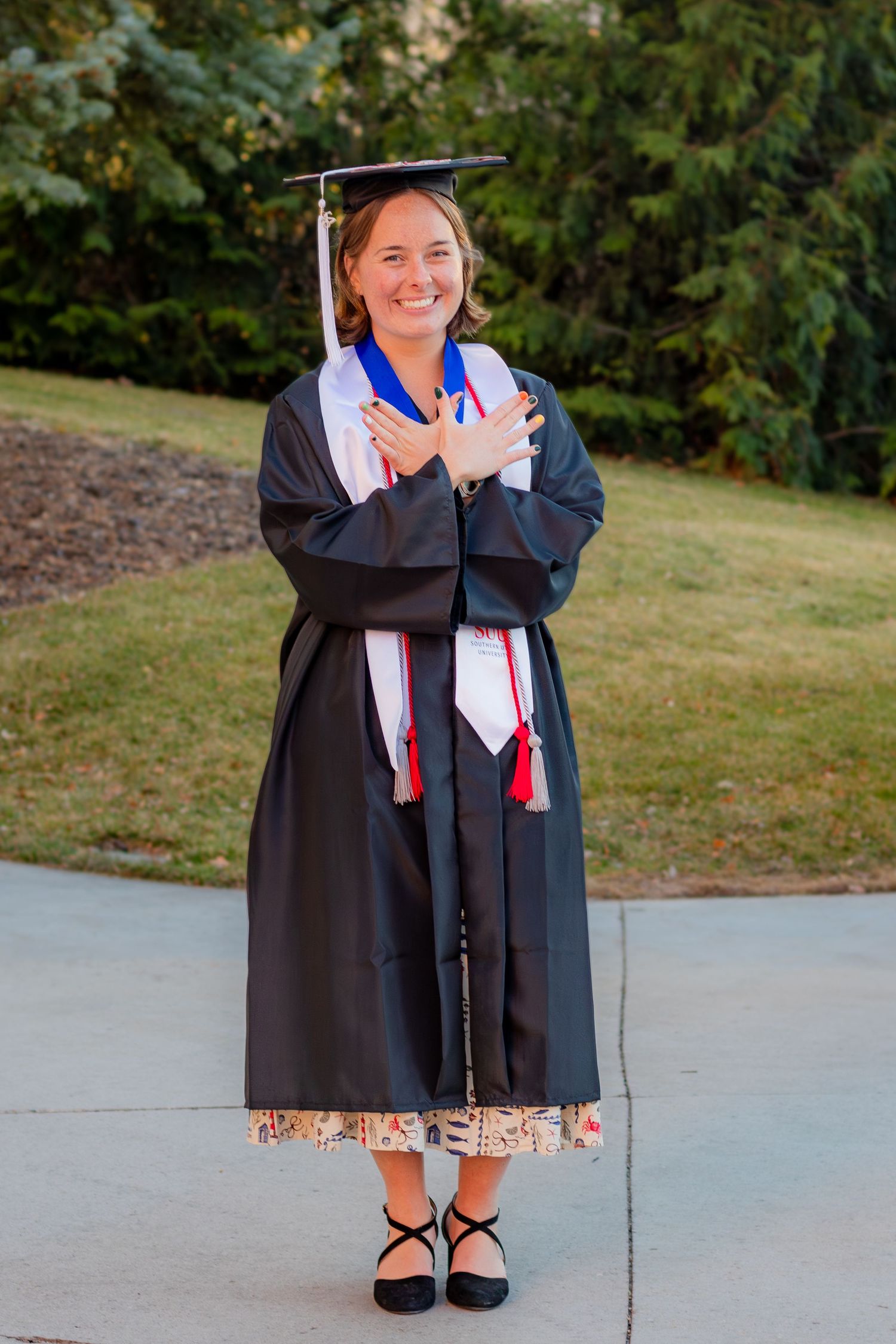 Smiling graduate in black cap and gown with colorful stoles poses outdoors on sunny day. Southern Utah University Senior Graduate
