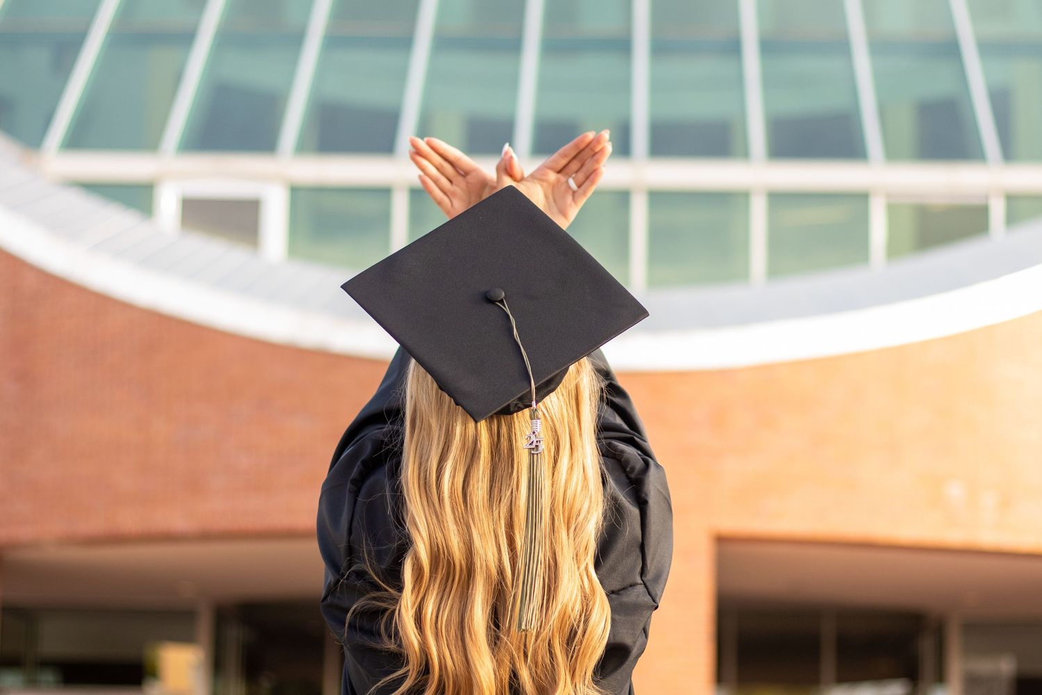 Graduate in cap and gown holds mortarboard hat, standing with back to camera outside modern university building.
