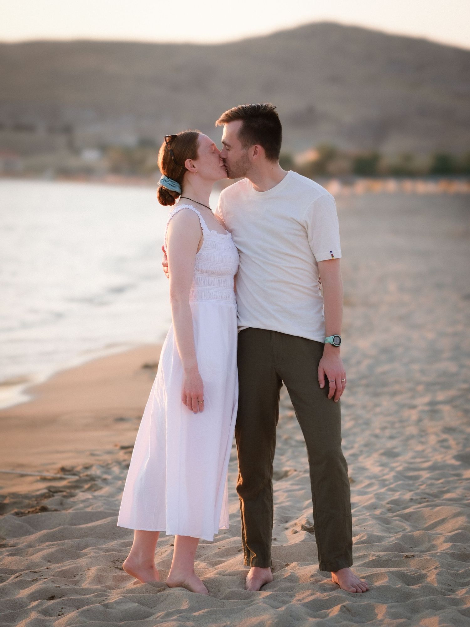 Barefoot couple kissing on beach at sunset with mountains in background.