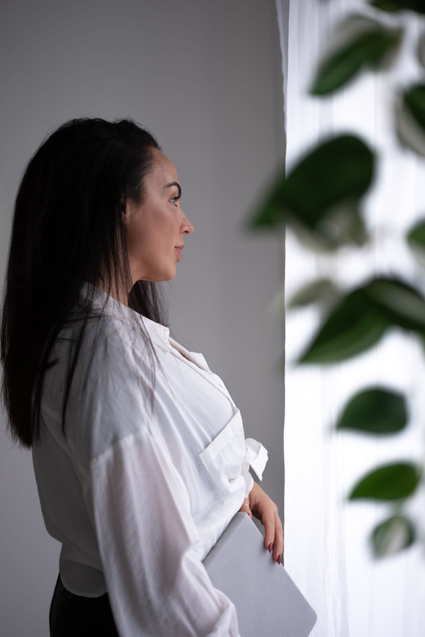 Elegant brunette woman in white blouse holding a laptop, gazing out a window near green plants.