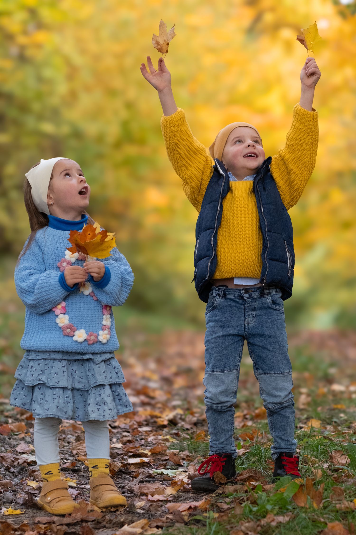 Two children in colorful autumn outfits toss yellow fall leaves in the air in a golden forest.