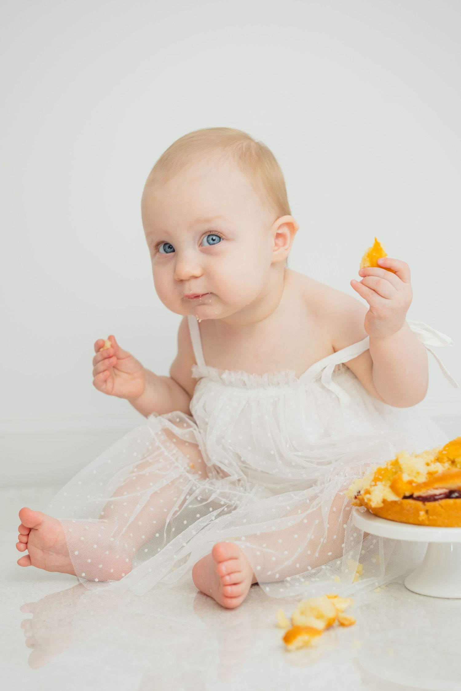 Baby girl in white dotted dress sits beside a smashed birthday cake, holding a piece of orange cake.