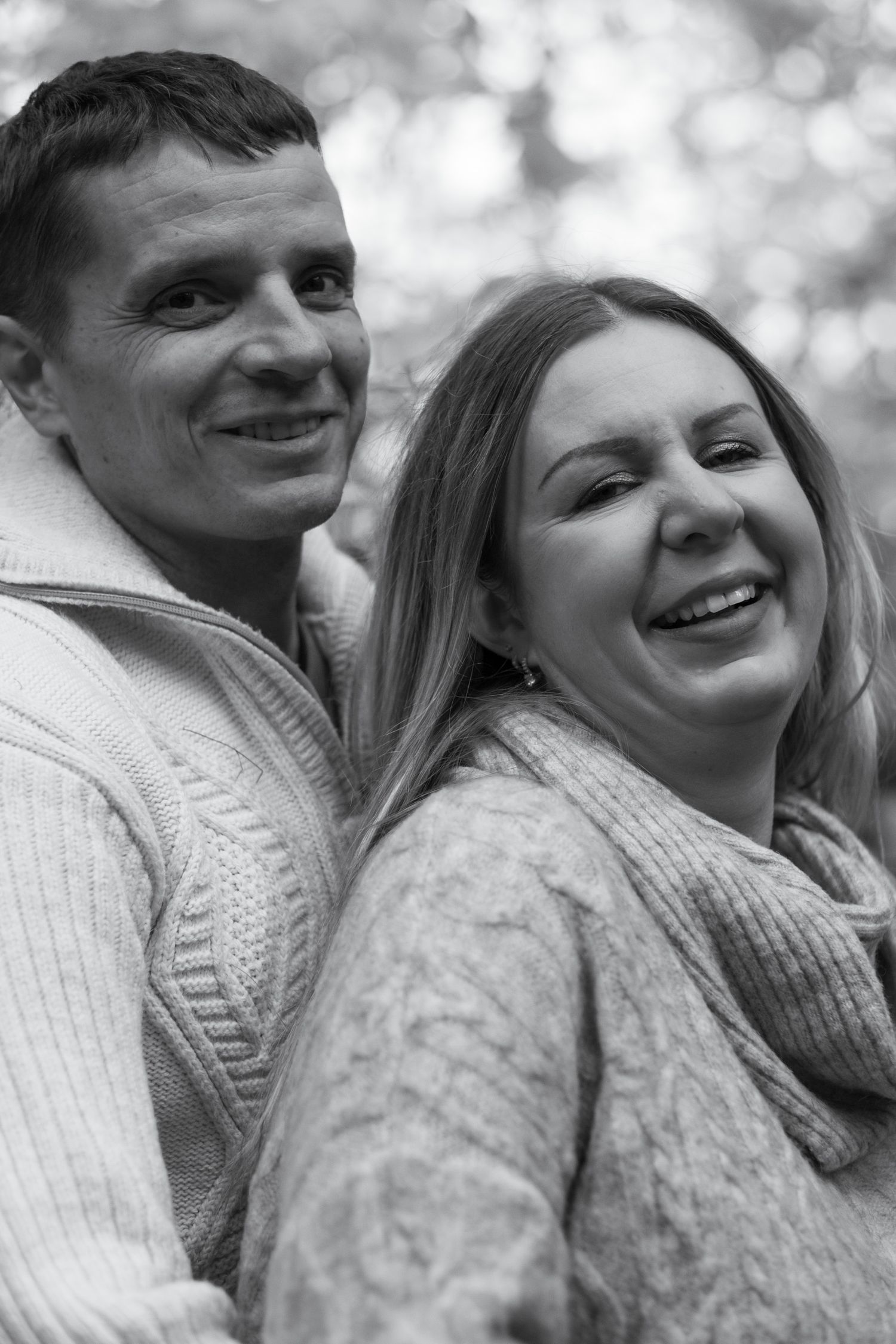 Black and white portrait of a smiling couple standing back to back outdoors in a park.