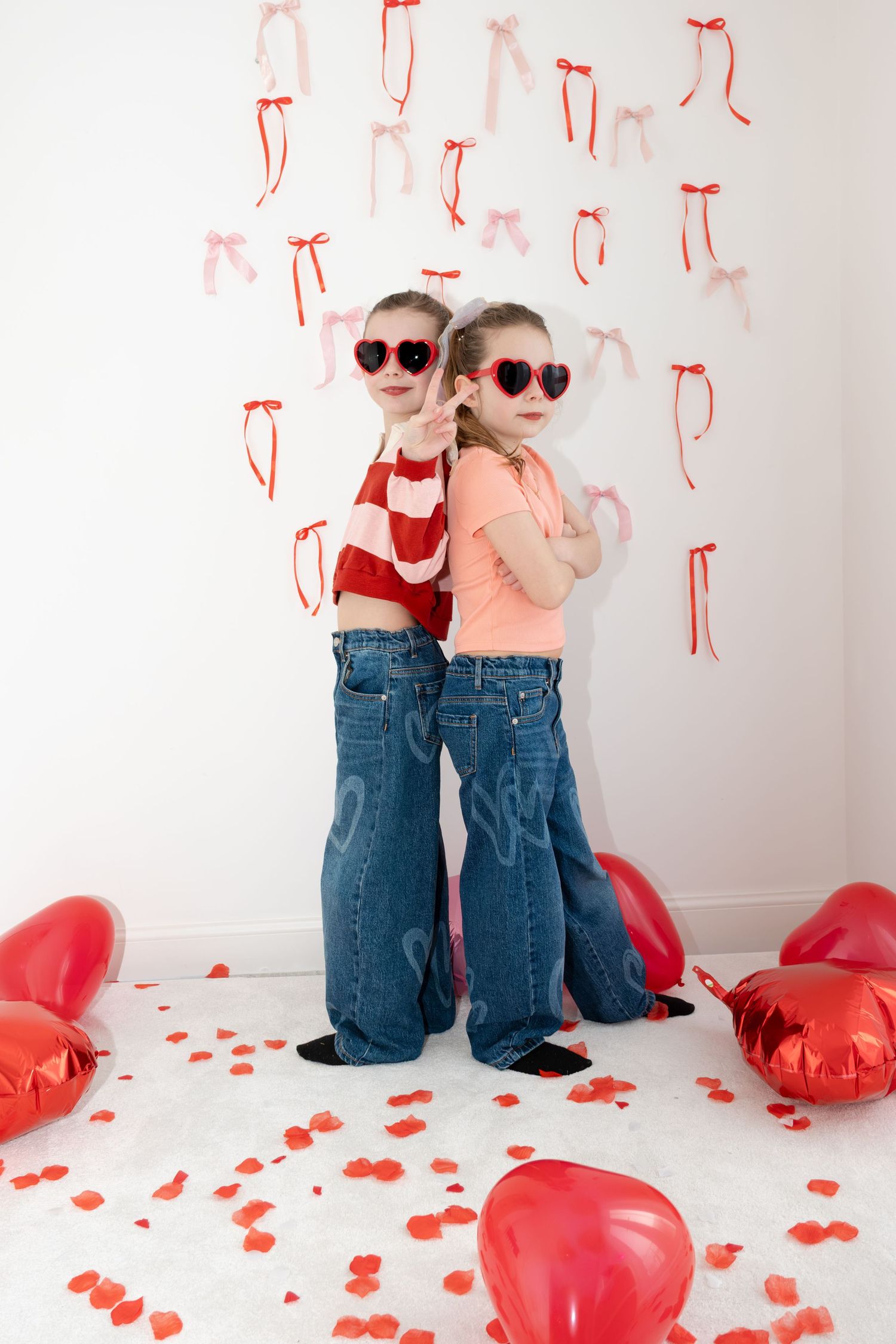 Two girls in jeans wearing heart sunglasses posing with red heart balloons and bows.