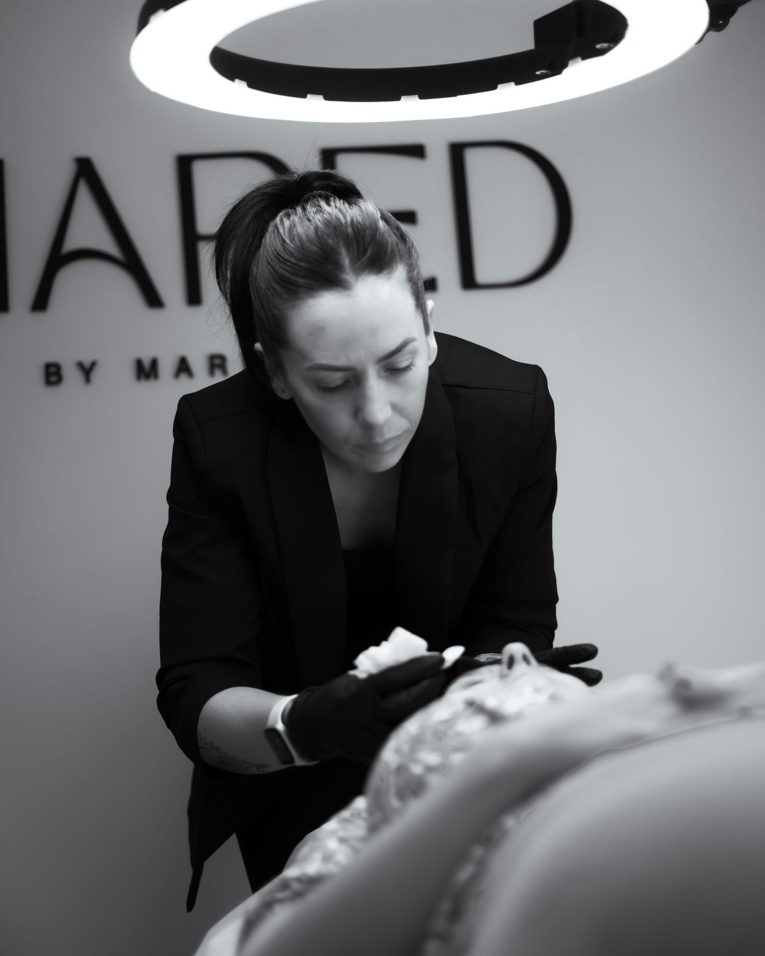 Black and white photo of a focused beauty professional performing a facial treatment under a ring light.