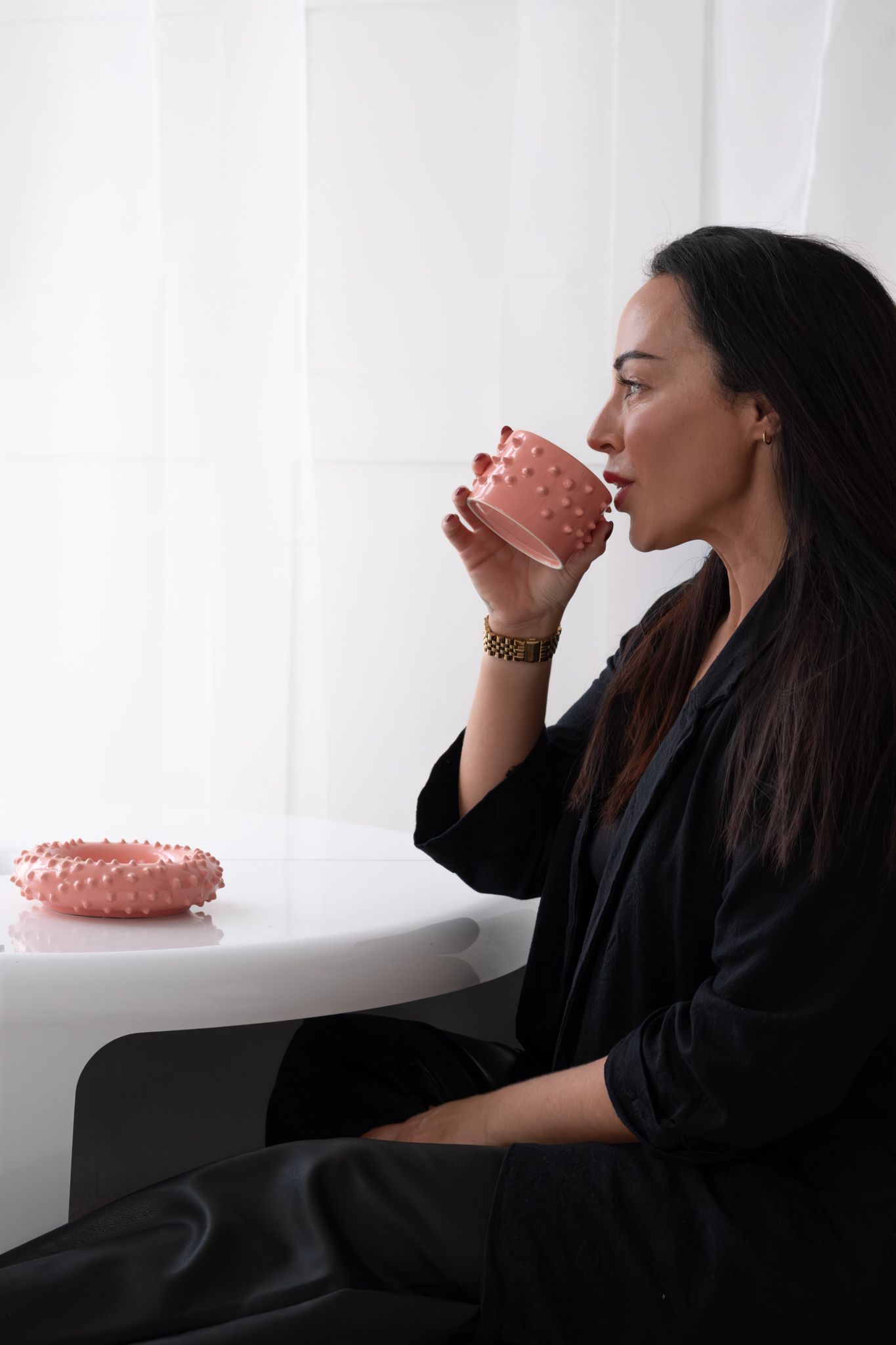 Elegant woman in black outfit sipping from pink textured mug at a small white round table.