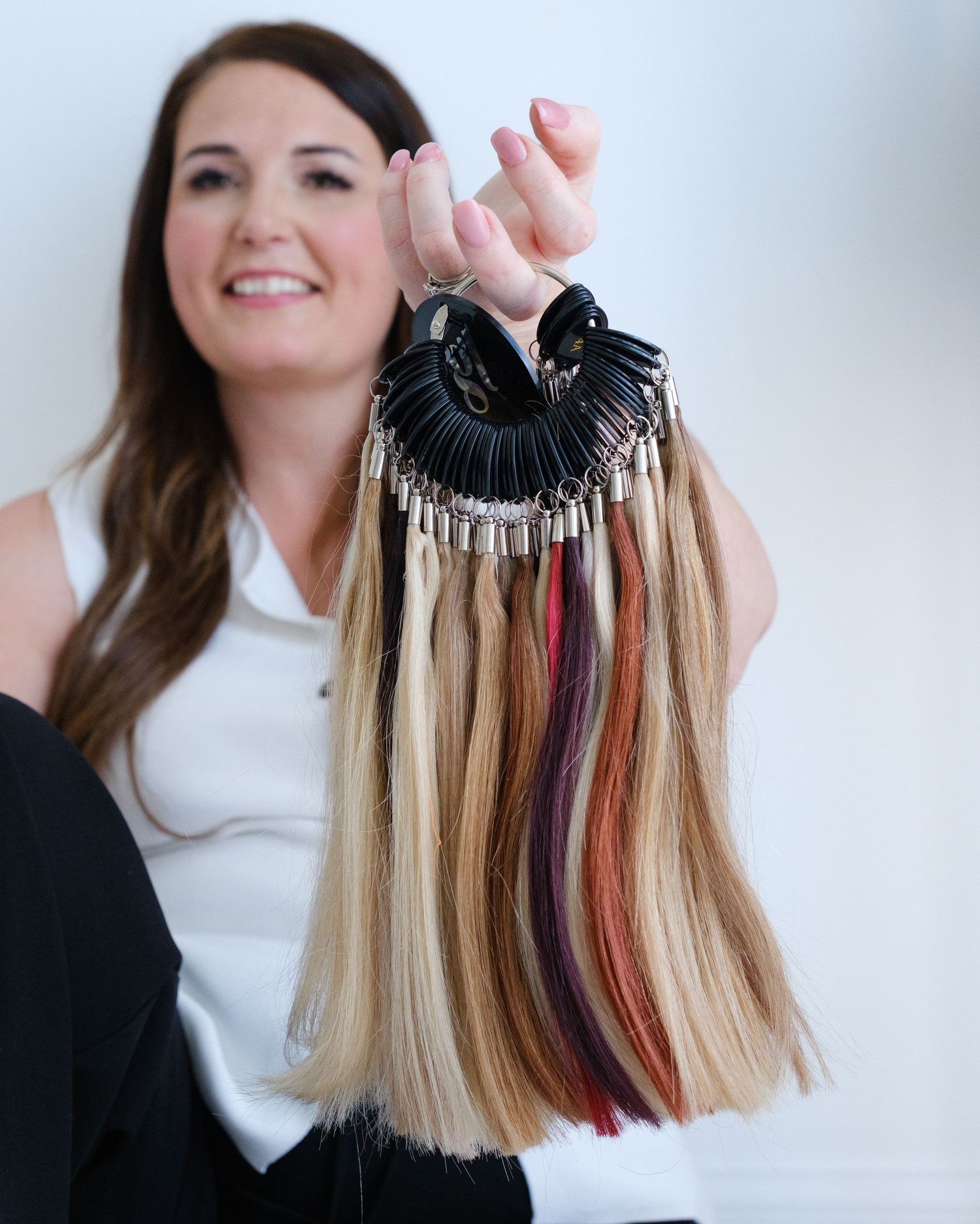 Smiling woman in white top holding a large hair color swatch ring with multiple shades and lengths.