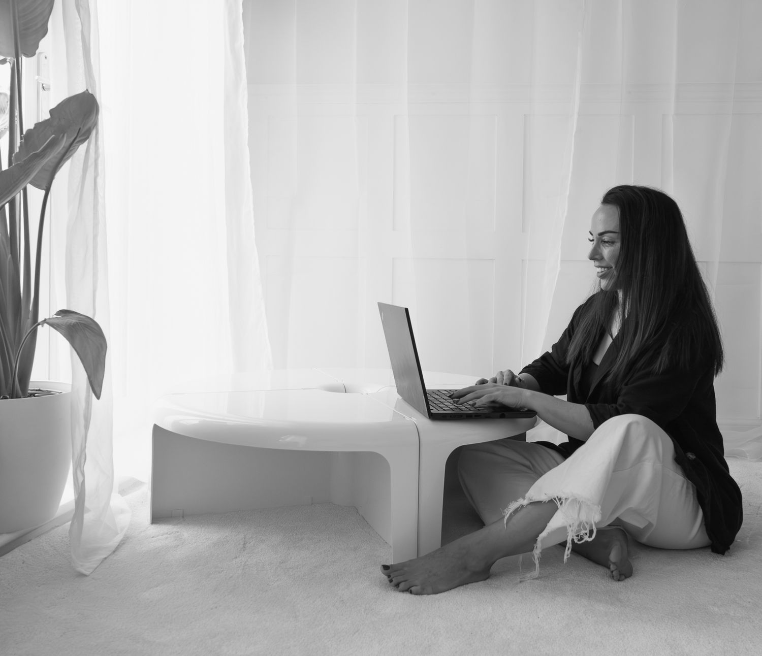 Black and white photo of a smiling woman sitting cross-legged on the floor working on a laptop.