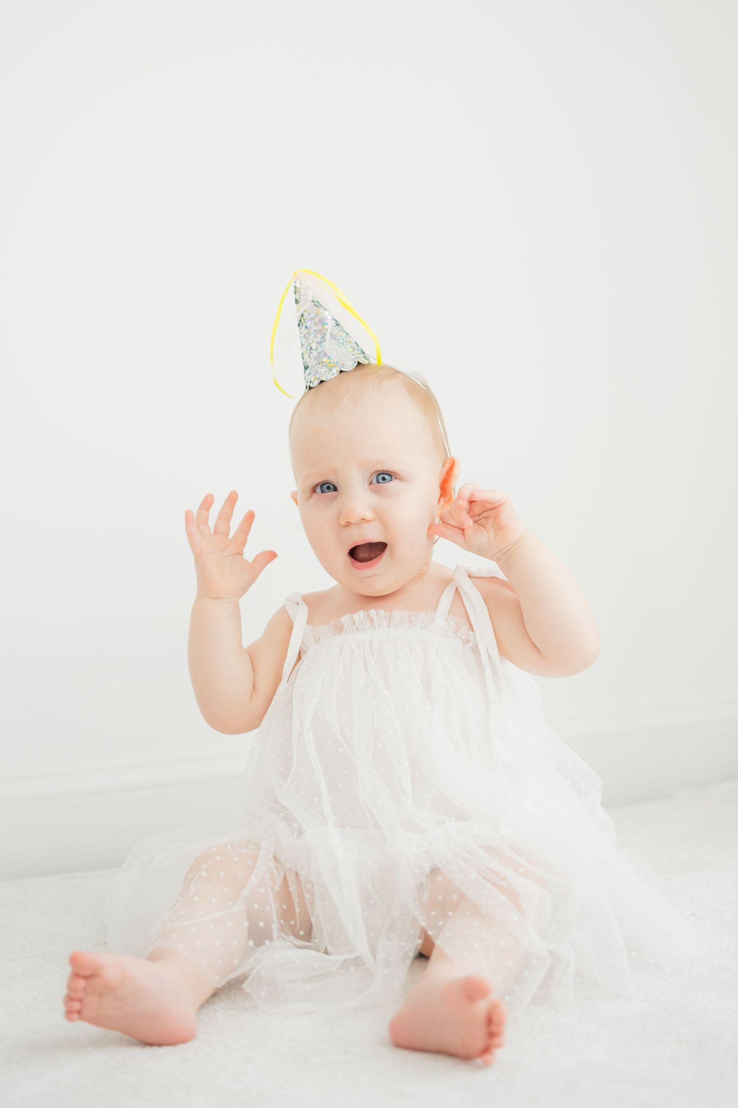 Excited baby girl in white dress and silver party hat claps hands while celebrating her first birthday.