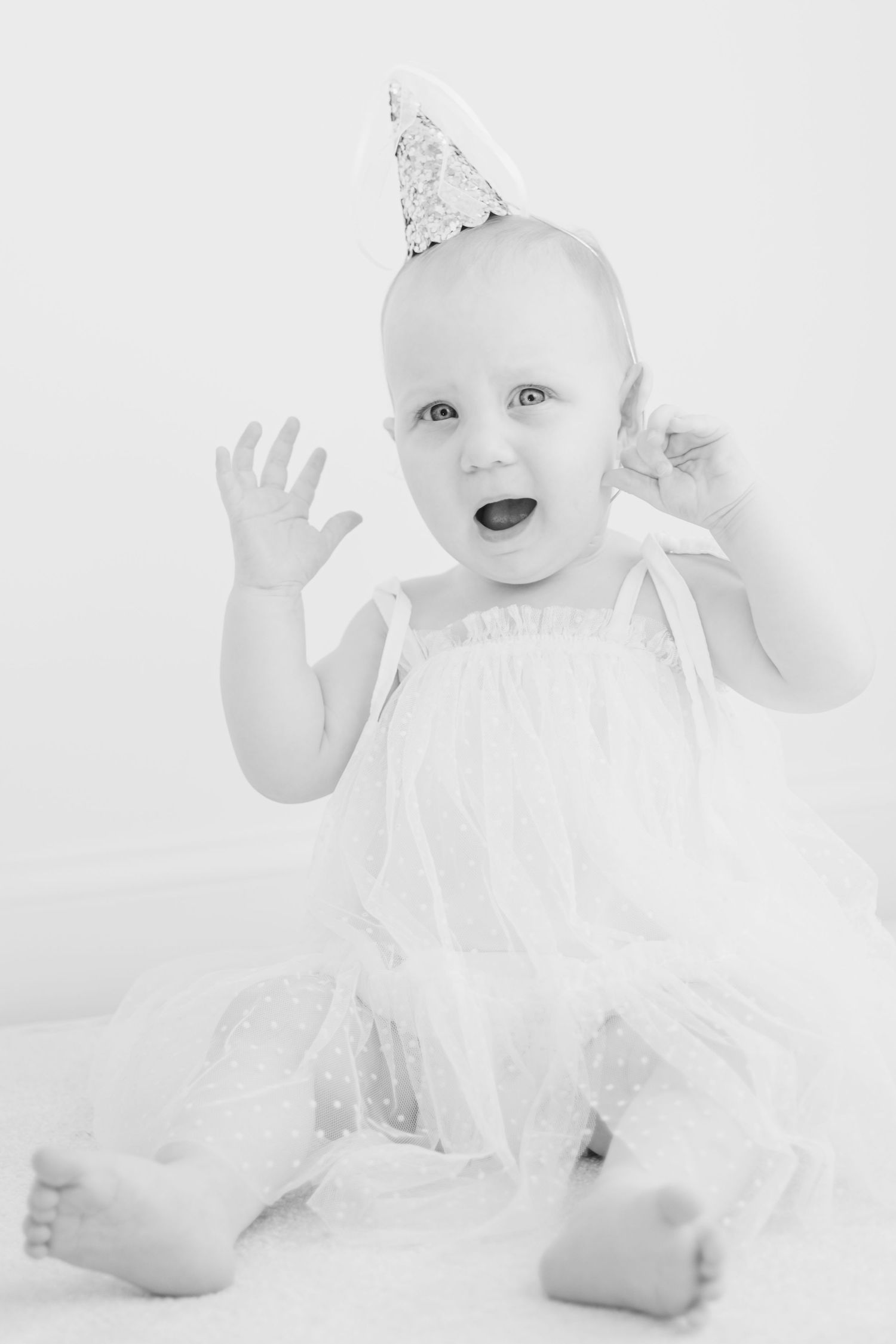 Black and white photo of a joyful baby in a white dress and party hat celebrating her first birthday.