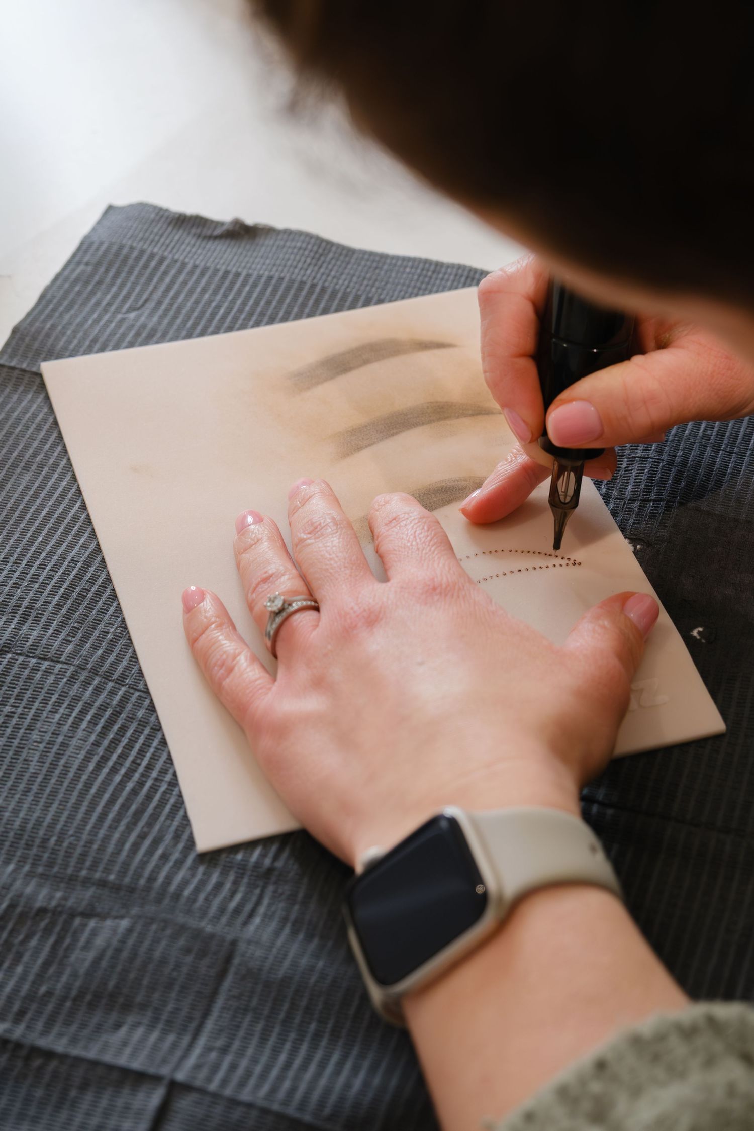 Close-up of hands practicing eyebrow mapping technique on a practice skin pad with a compass tool.