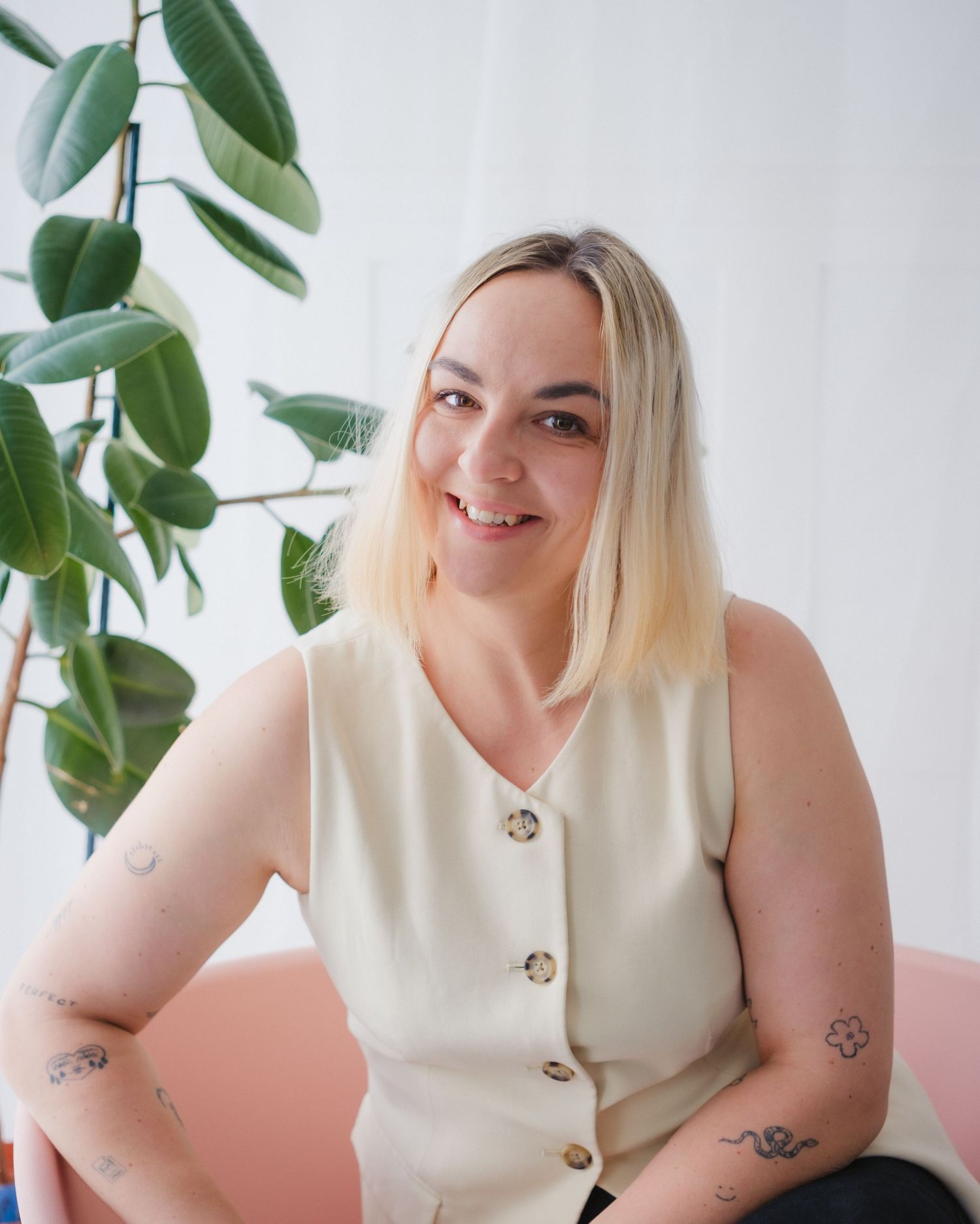 Smiling blonde woman with tattoos wearing cream sleeveless vest sitting near green indoor plant.