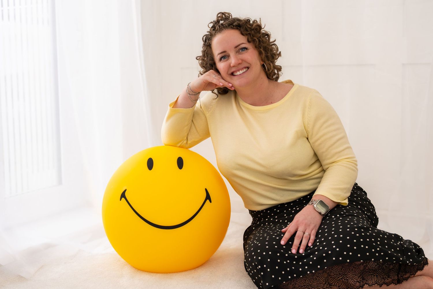 Smiling curly-haired woman sitting beside large yellow smiley face balloon on white floor.
