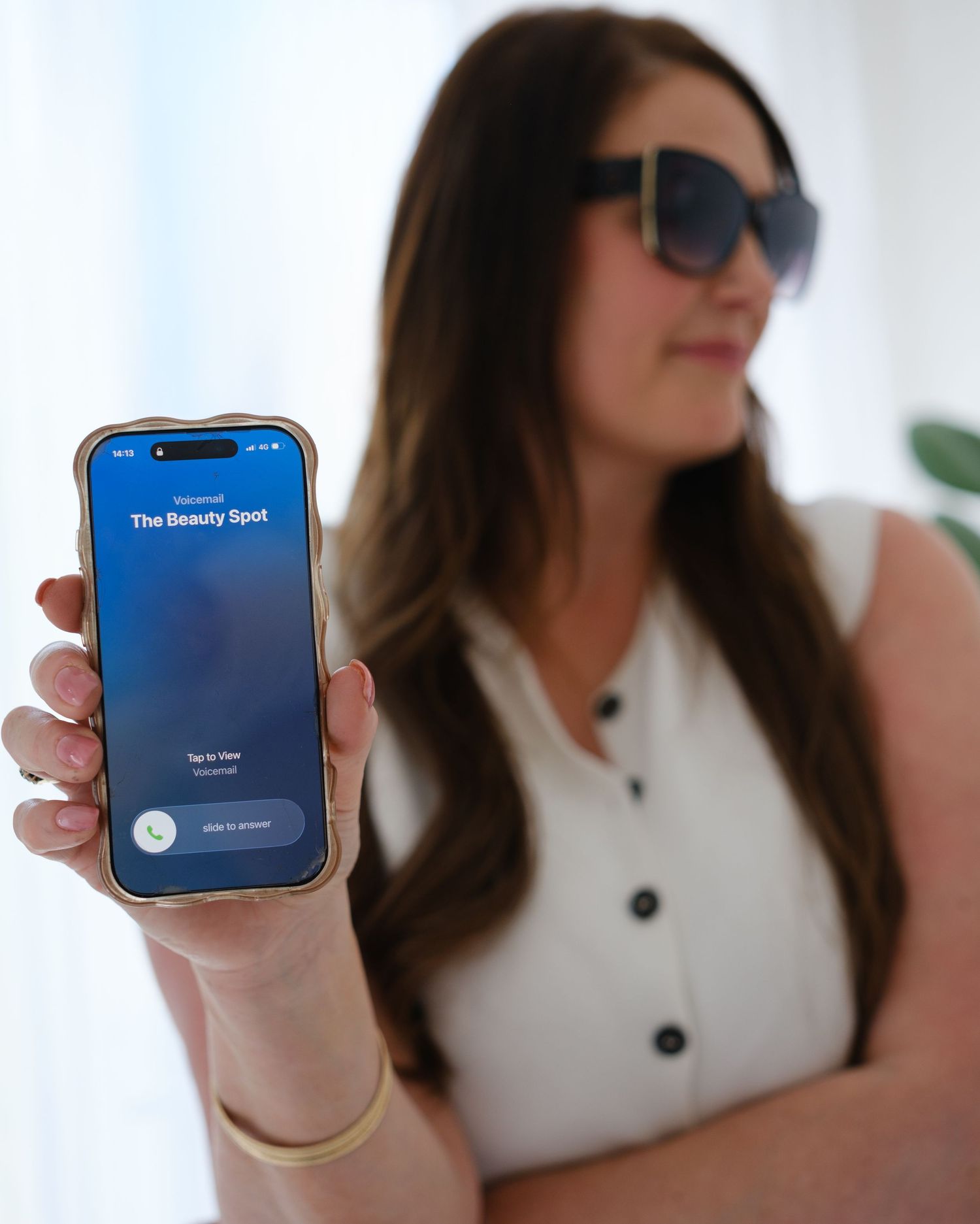 Woman in sunglasses holding an iPhone displaying The Beauty Spot app screen in a bright studio setting.