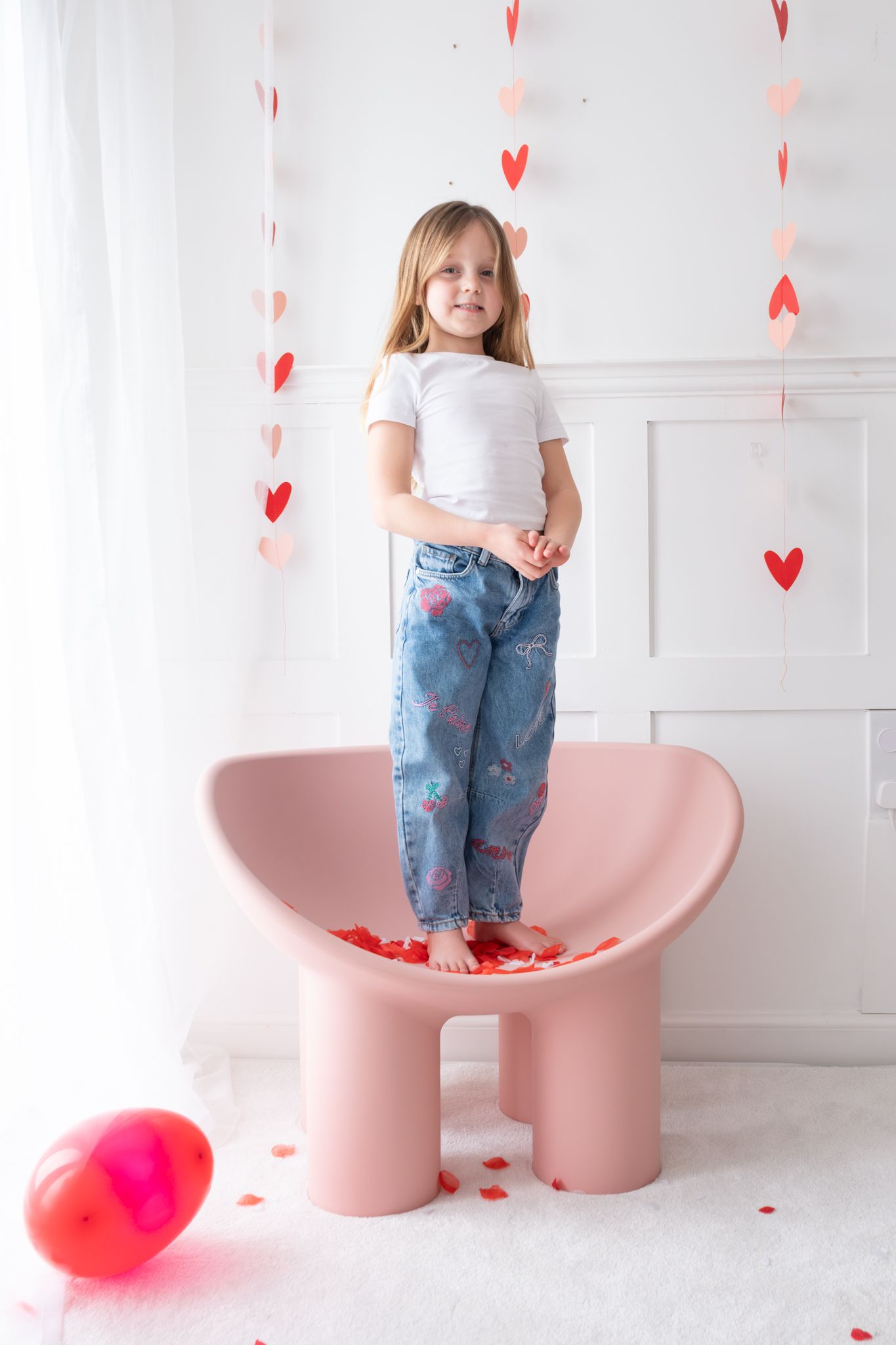 Young girl in white top and jeans standing on a pink chair with Valentine heart decorations.