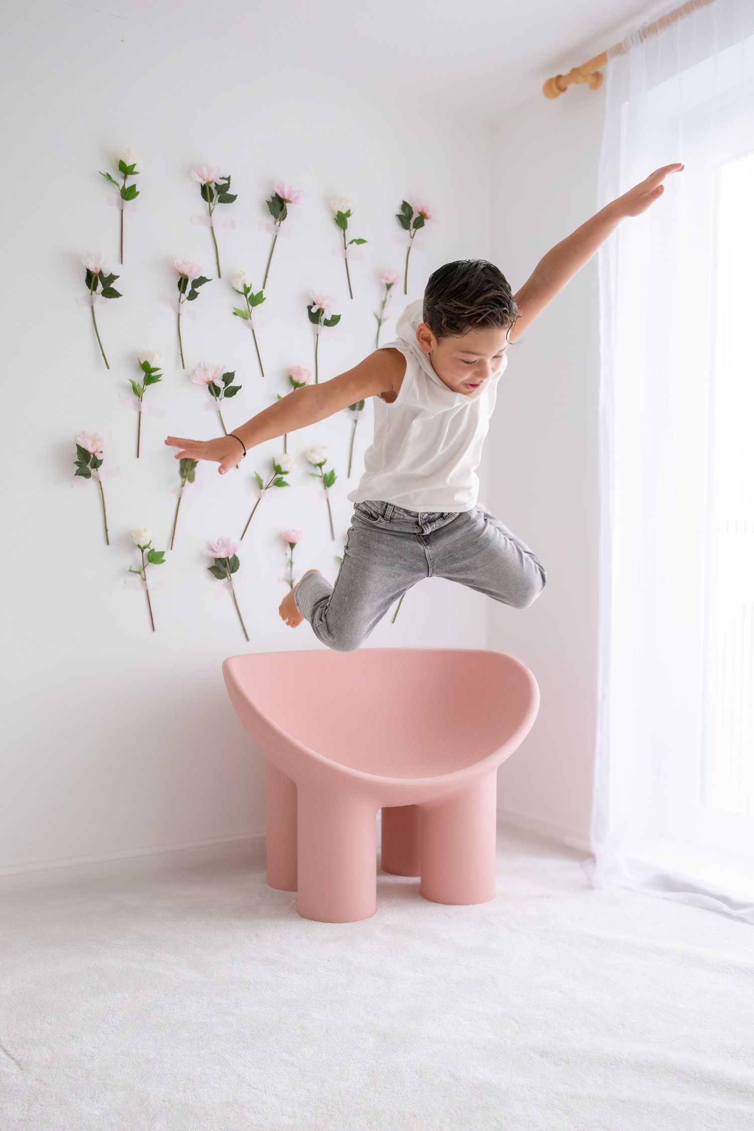 Boy in white tank top jumping energetically near pink chair with flowers on white wall.