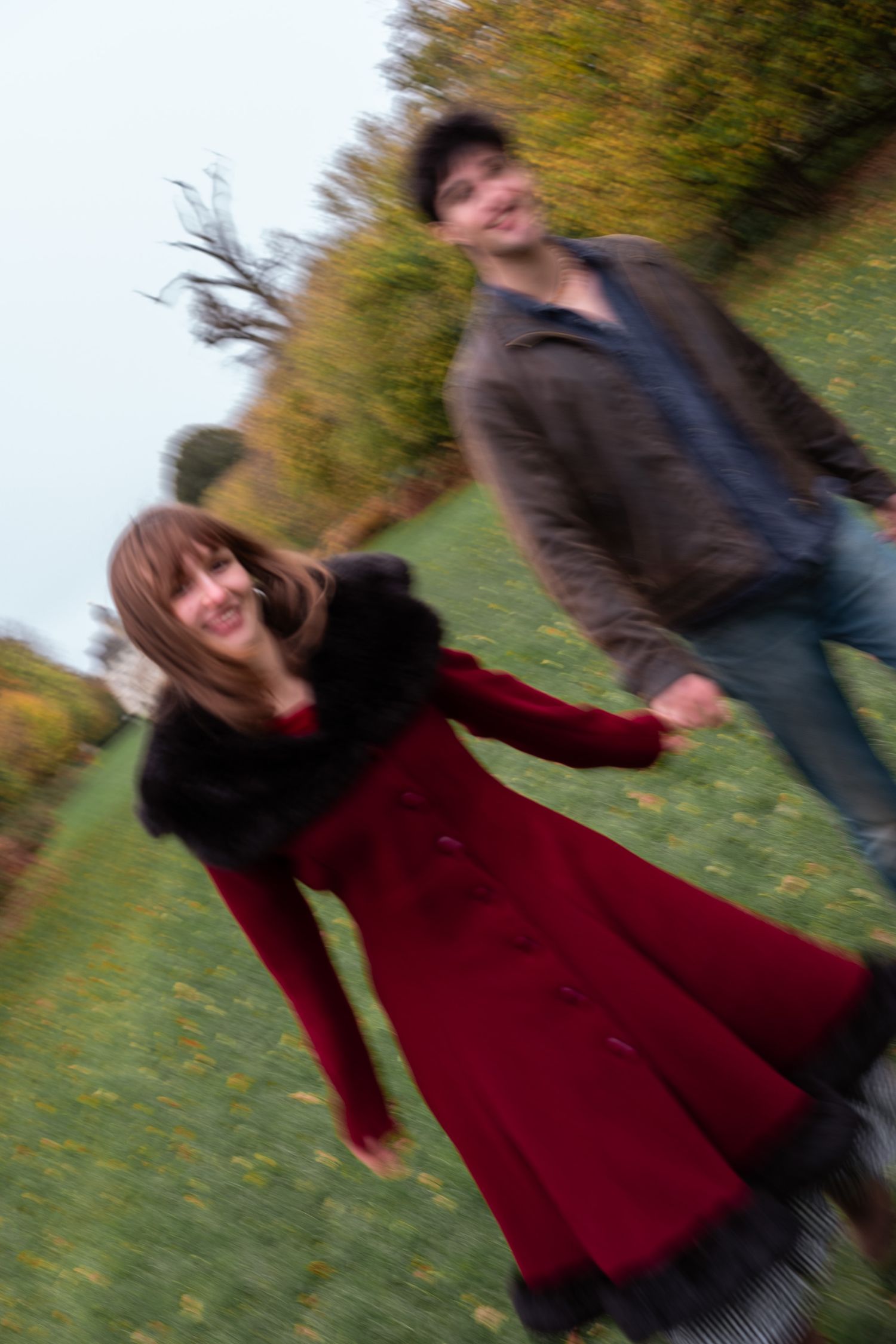 Blurred motion shot of happy couple holding hands running through autumn park, woman in red coat laughing.