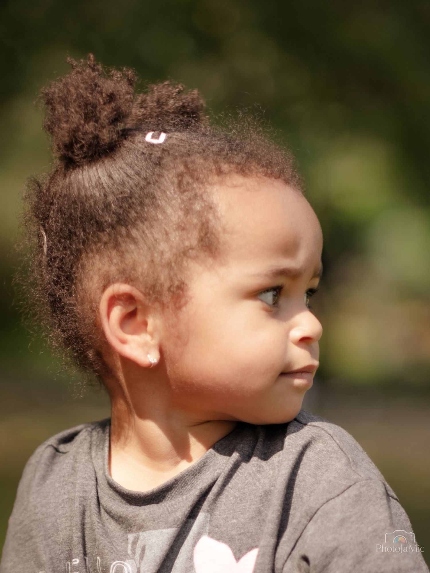 Profile portrait of young girl with curly hair bun wearing gray shirt outdoors.