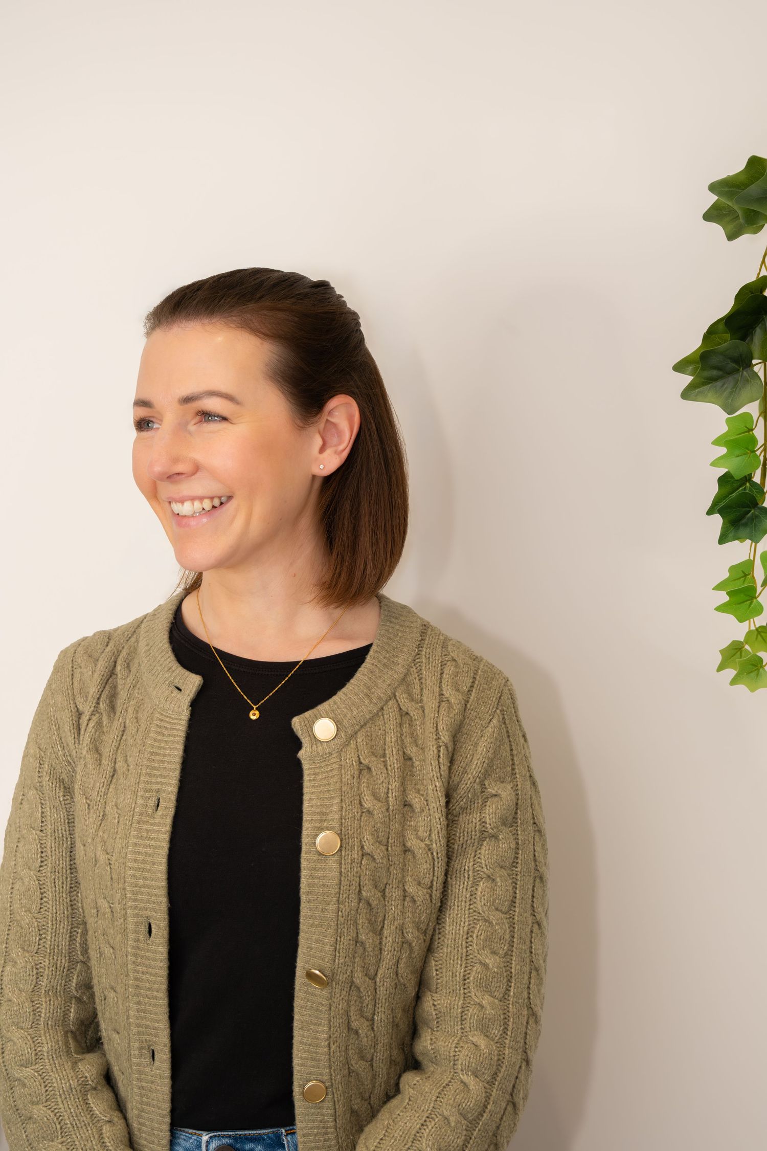 Smiling woman in sage green cable knit cardigan stands against white wall beside green ivy plant.