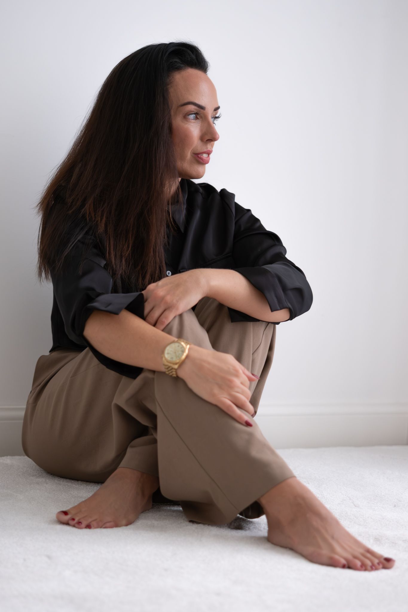 Barefoot woman in black shirt and tan pants sits on floor gazing sideways in bright white room.