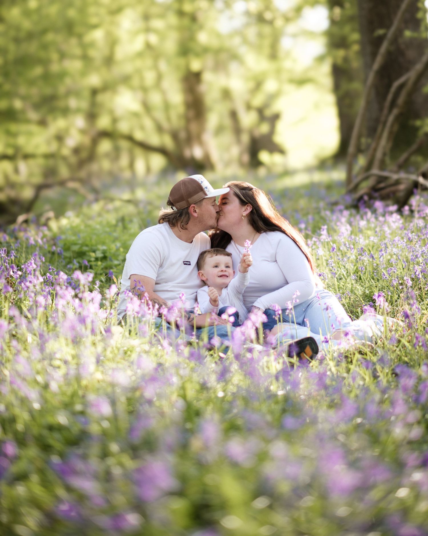 Couple kissing with baby in field of purple wildflowers in sunlit woodland setting.